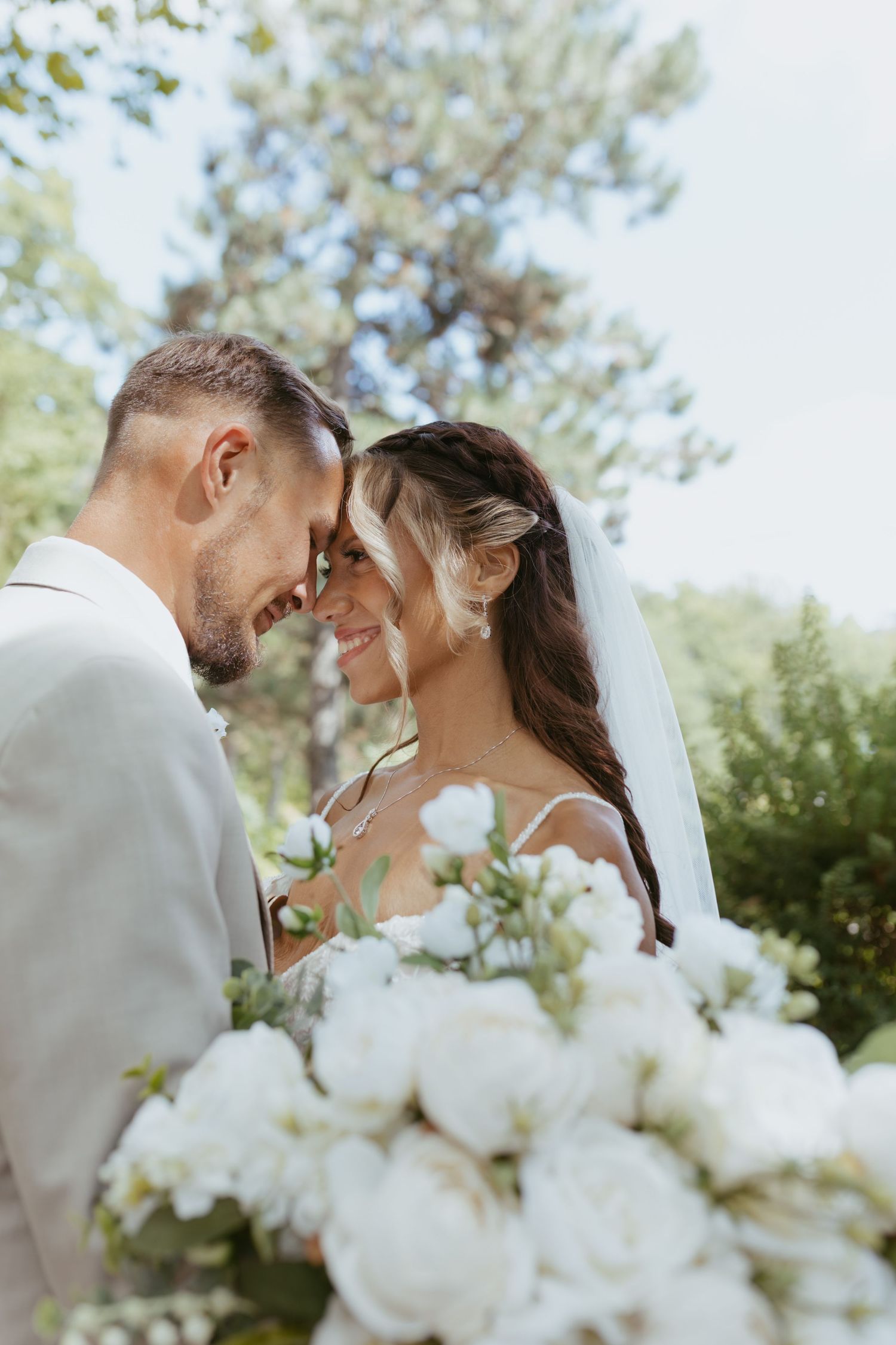 Bride and groom touching foreheads, smiling, with white peony bouquet outdoors.