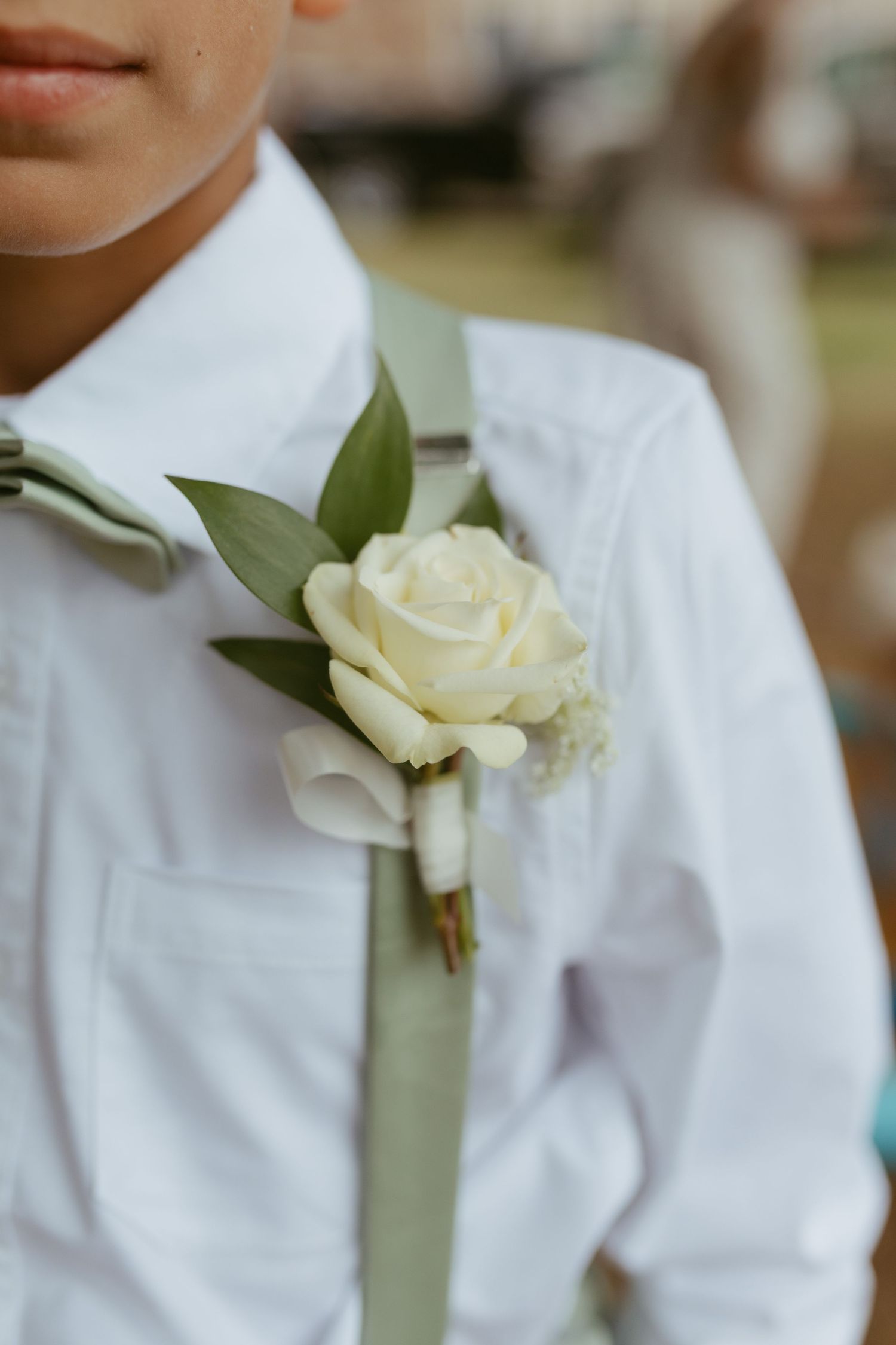 Close-up of a white rose boutonniere with green leaves on a boy's white shirt and sage green tie at a wedding.