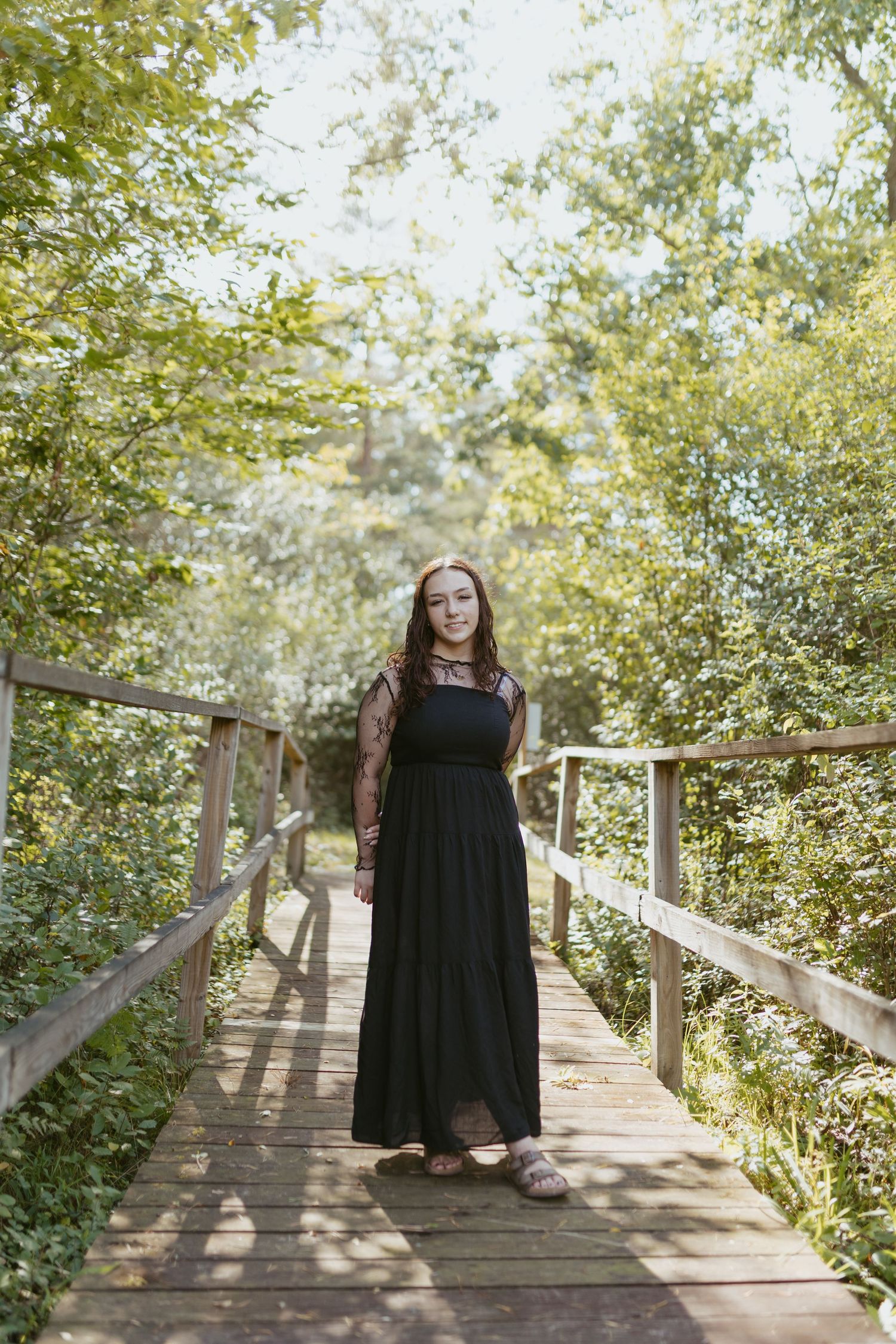 Young woman in black maxi dress standing on wooden bridge surrounded by lush green trees in sunlit forest.