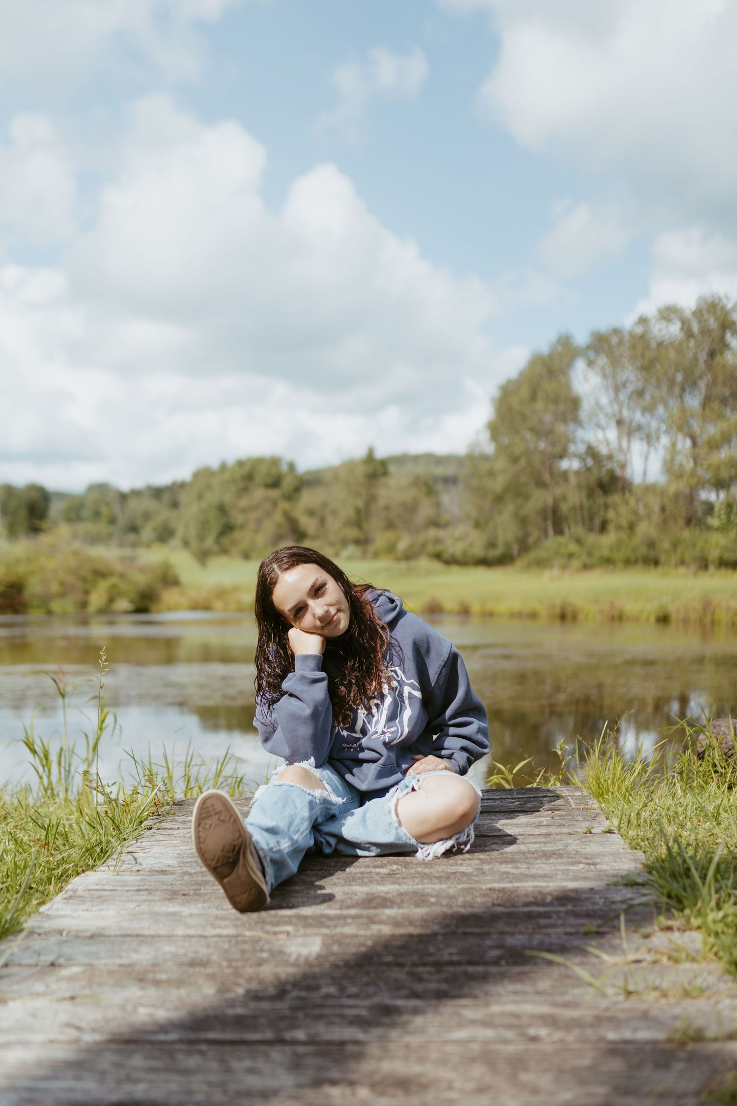 Young woman in hoodie and ripped jeans sitting on wooden dock by a serene lake on a sunny day.