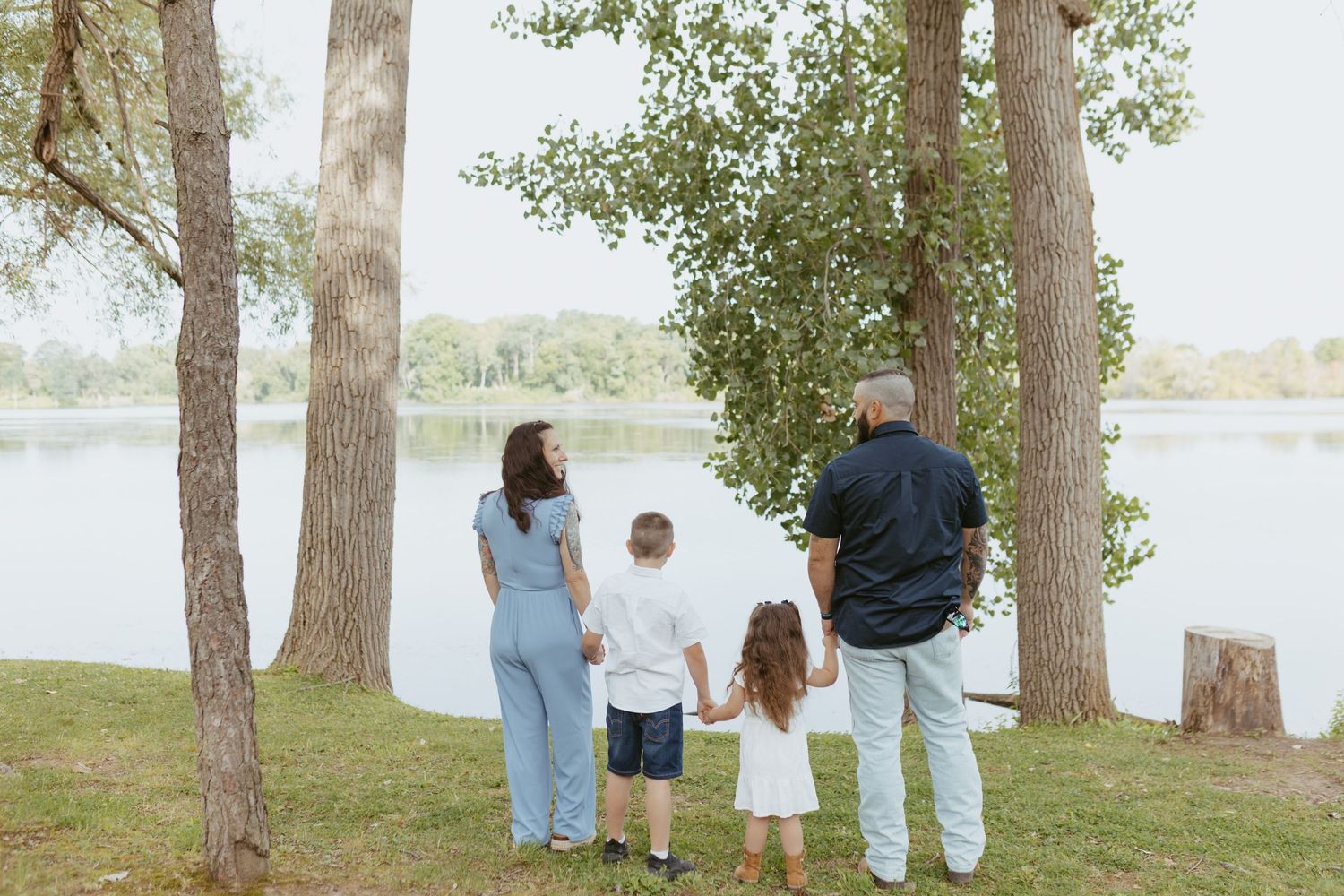 Family of four holding hands by a lakeside park with tall trees on a sunny day.