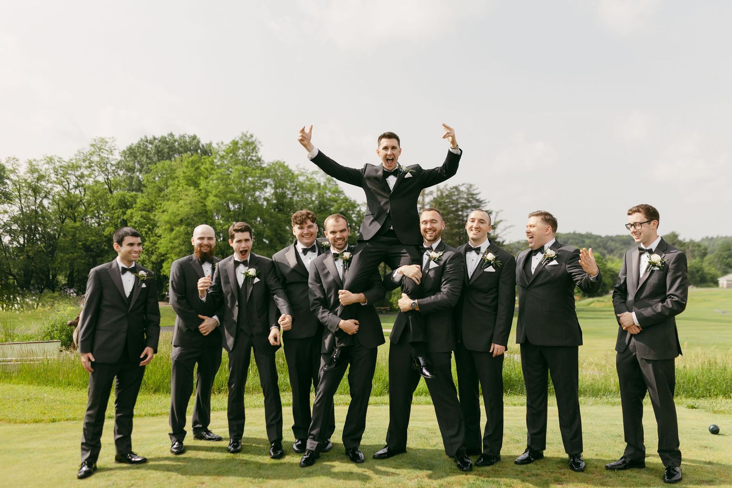 Groomsmen in black suits pose with celebratory gestures on a golf course.