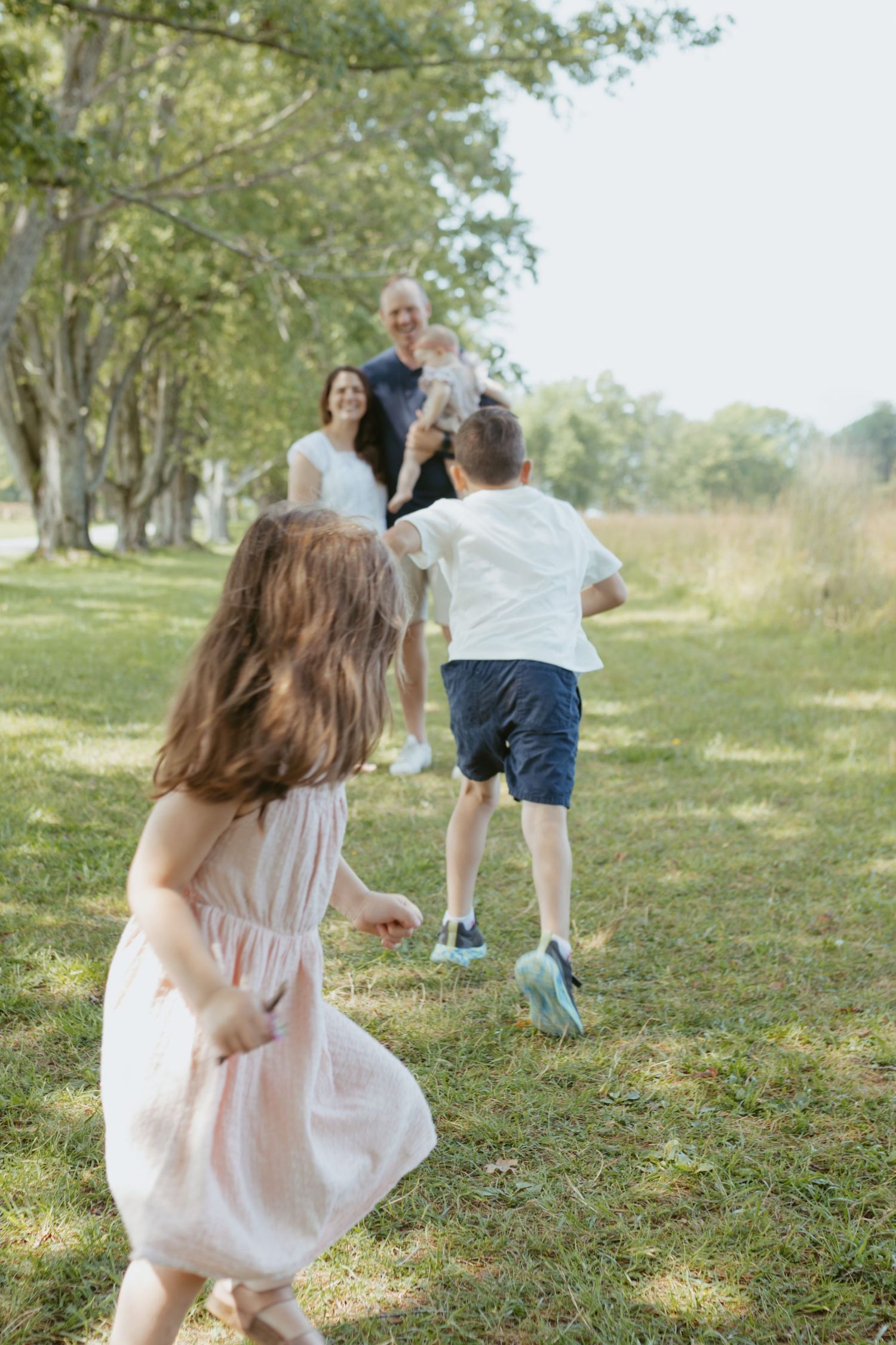 Children running toward parents in a sunny park, family playing outdoors on green grass with trees in background.