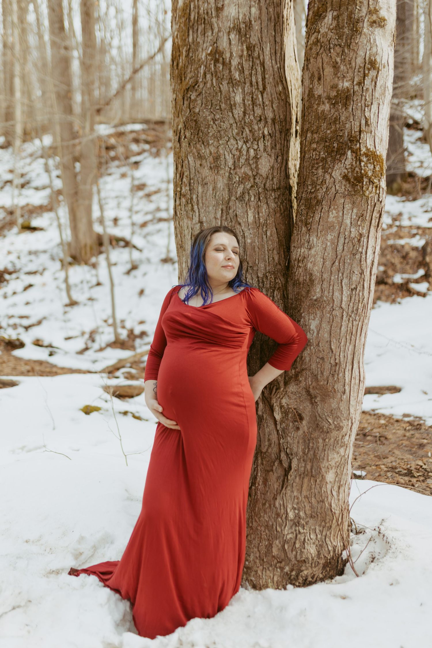 Pregnant woman in red gown leaning against tree in snowy forest during winter maternity photo shoot.