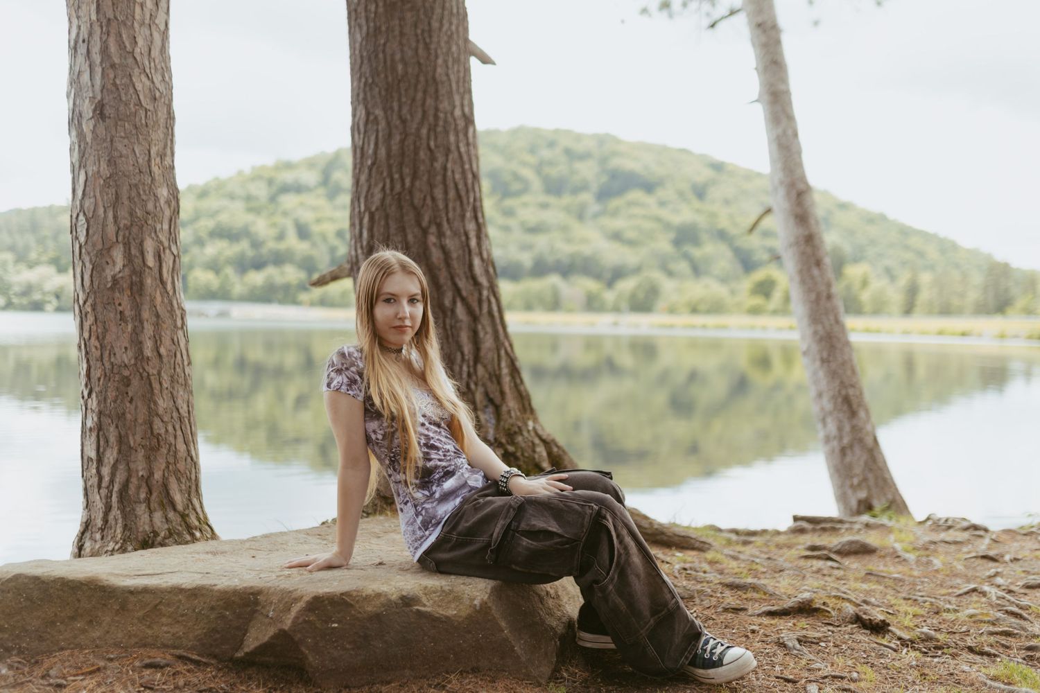 Young woman with long blonde hair sitting on a rock by a serene lake surrounded by tall trees and green hills.