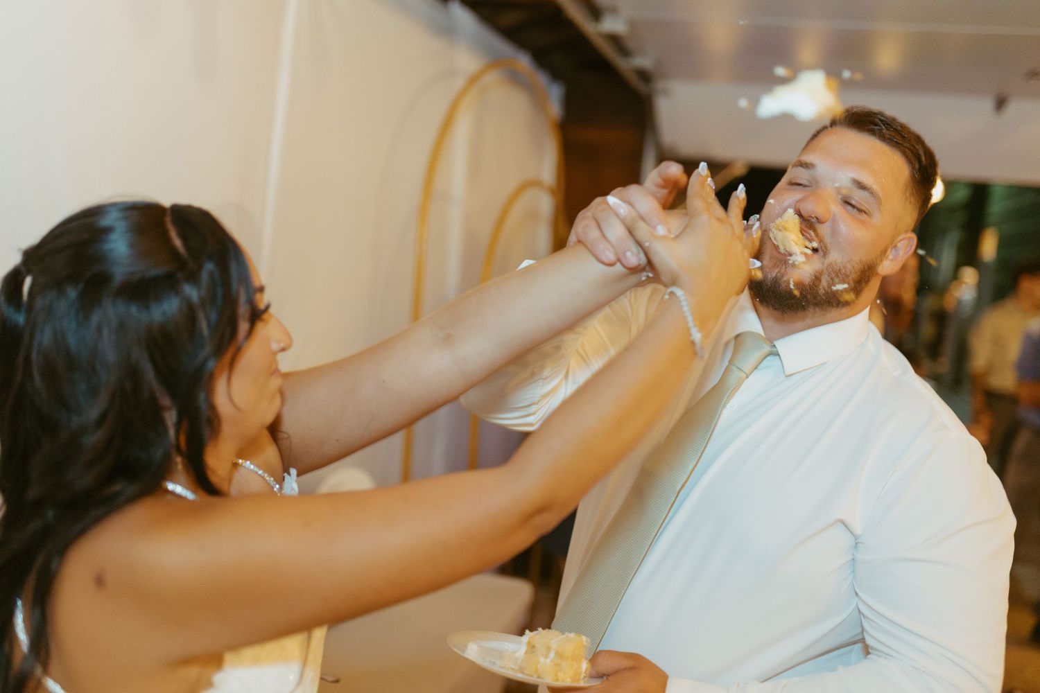 Bride smearing wedding cake on groom's face during fun cake cutting ceremony at reception.