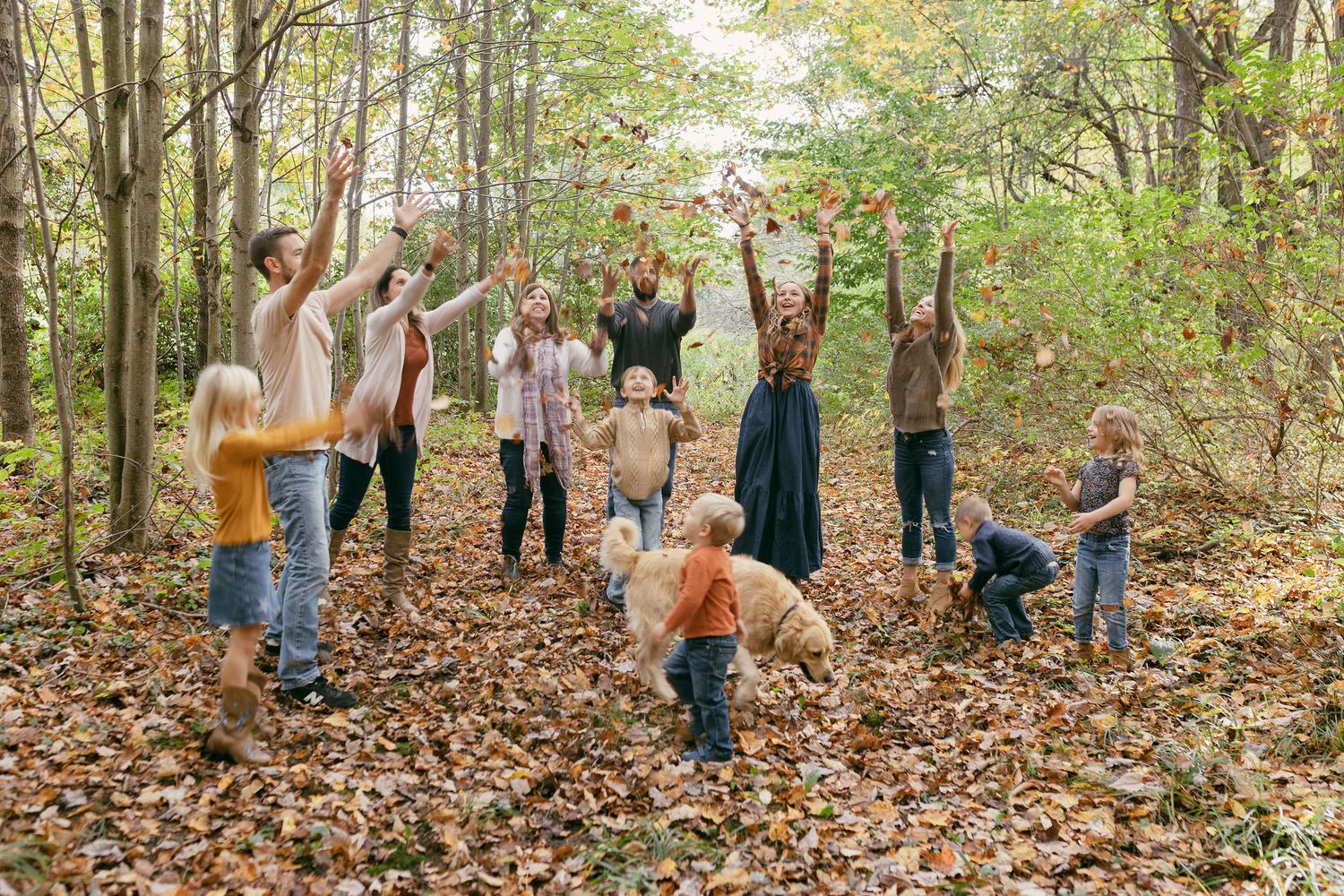 A joyful group of adults and children tossing autumn leaves in a forest clearing surrounded by fall foliage.