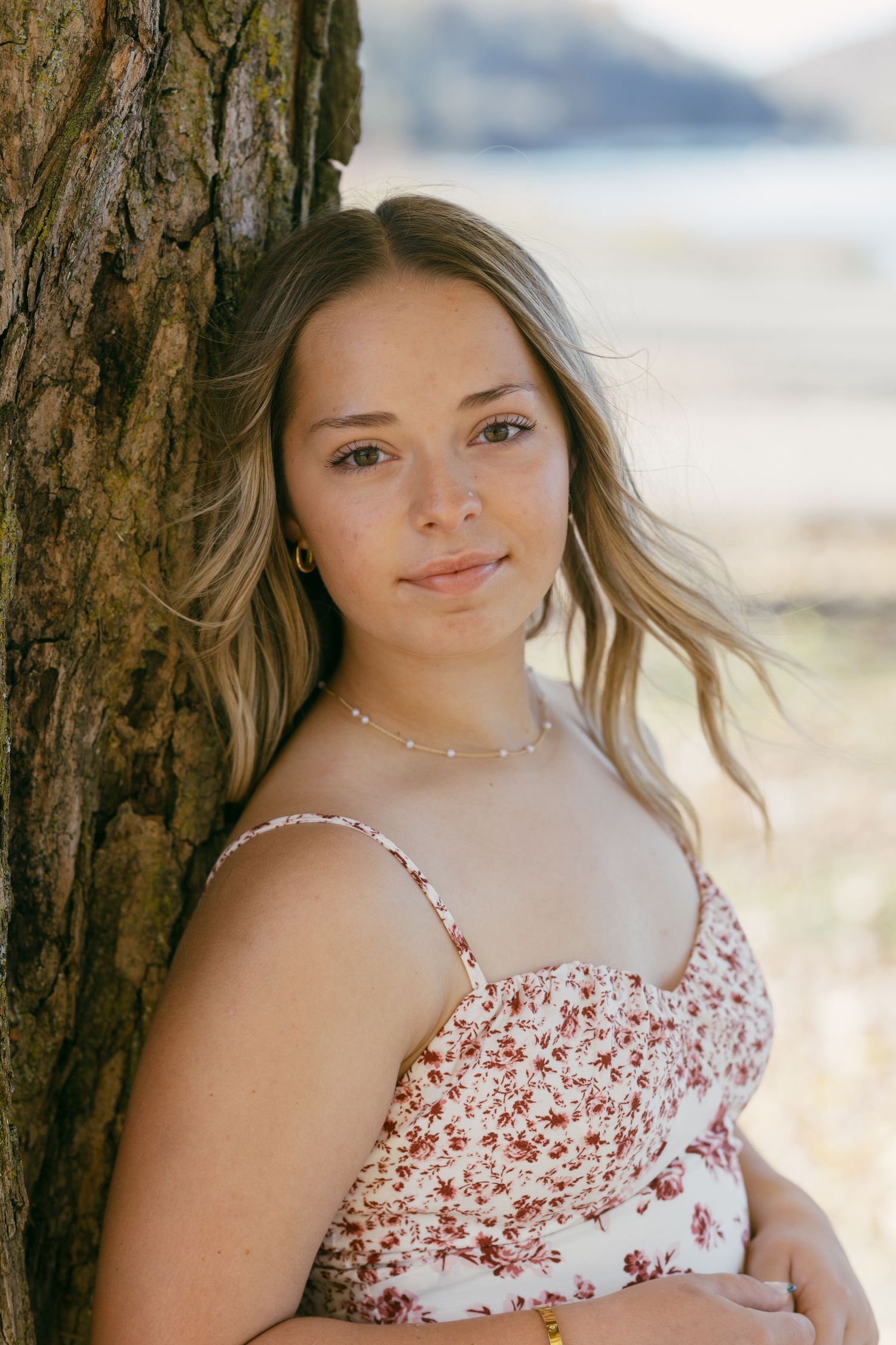 Young woman in floral sundress leaning against tree trunk outdoors, with soft natural background lighting.