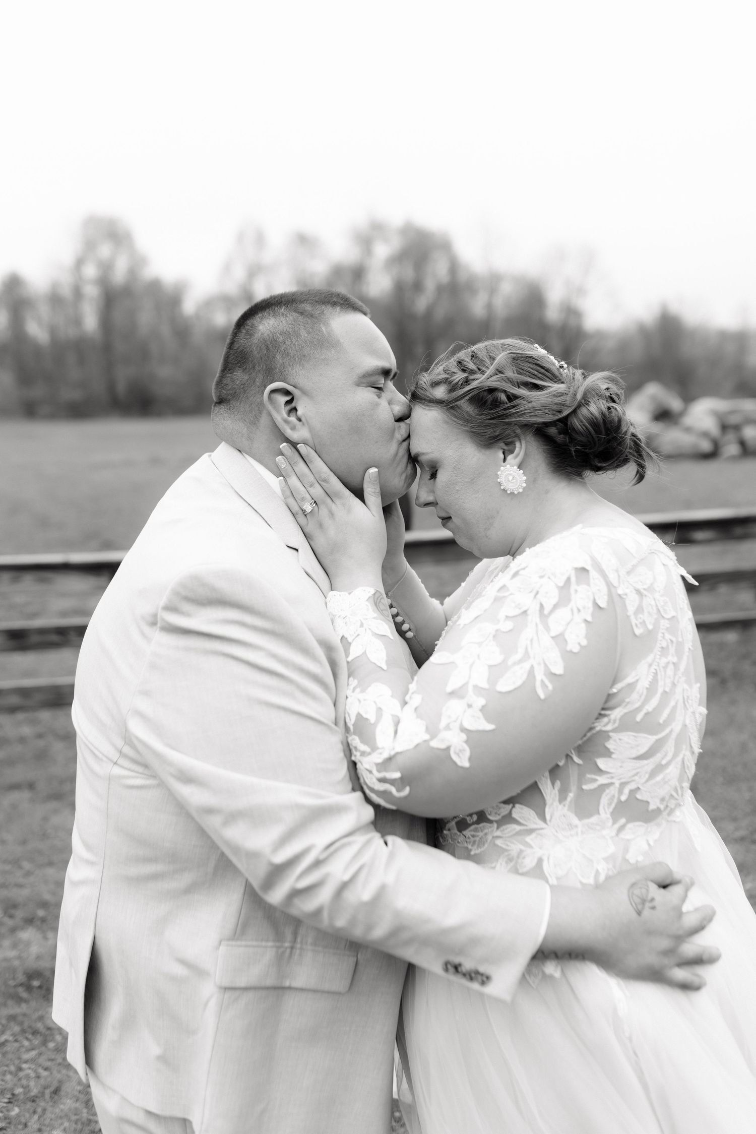 Black and white photo of groom kissing bride on forehead during romantic outdoor wedding portrait.