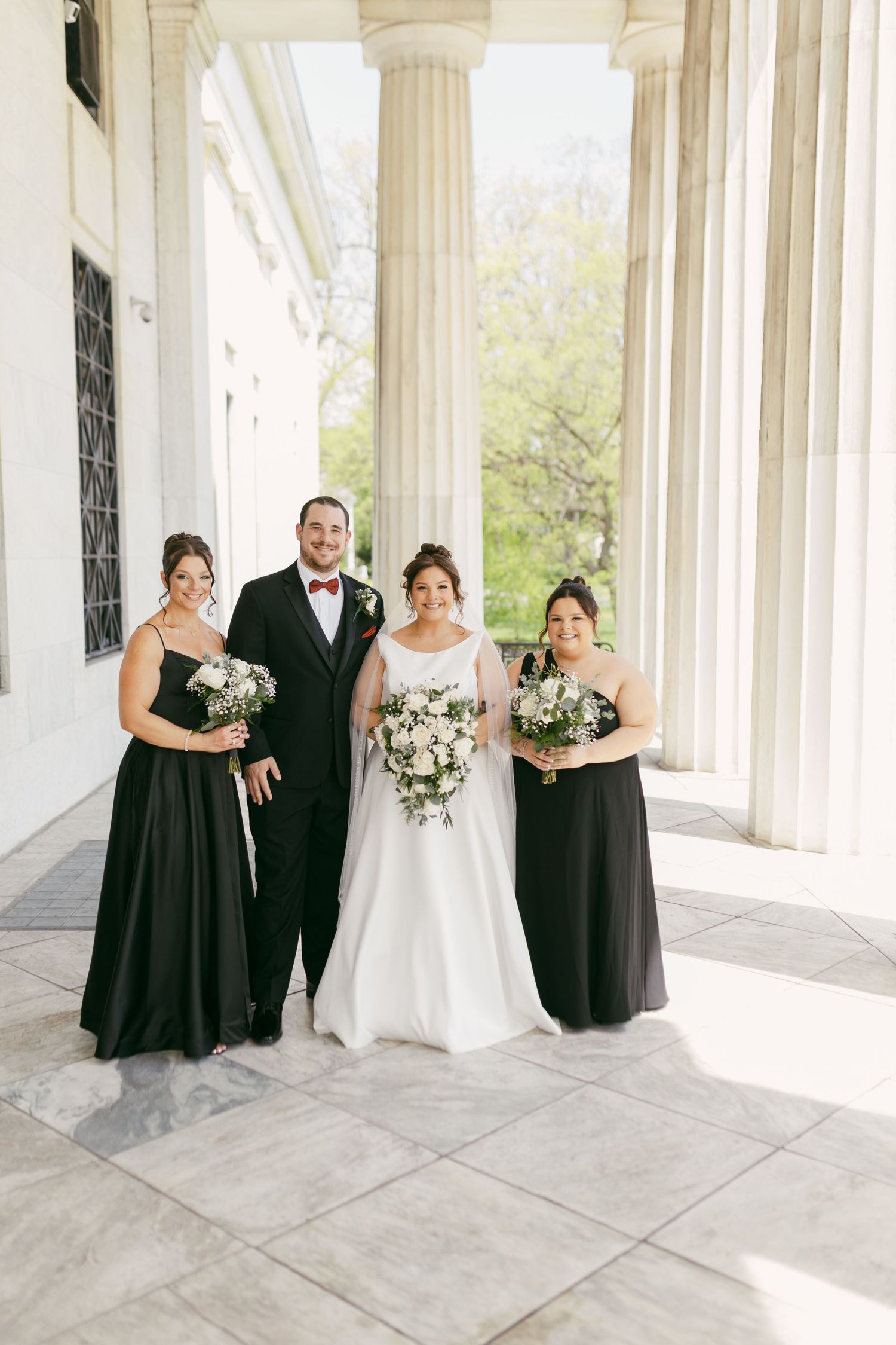 Bride and groom with two bridesmaids in black gowns posing between marble columns at wedding venue.