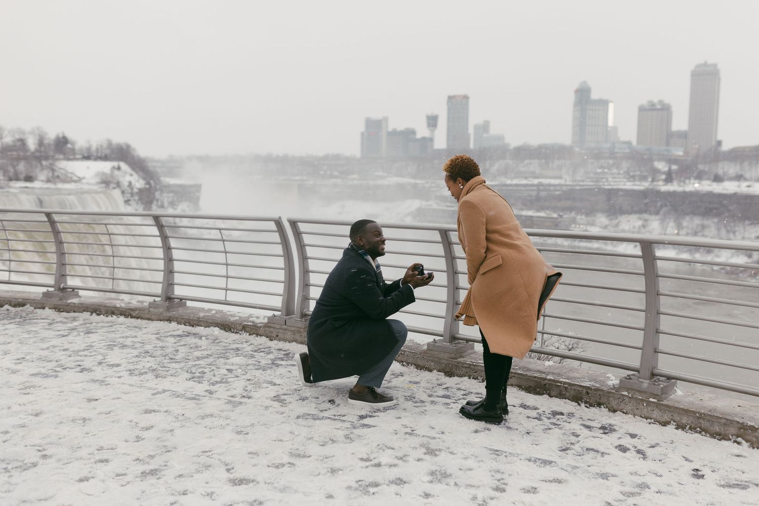 Man proposing to woman on snowy waterfront with city skyline backdrop in winter.