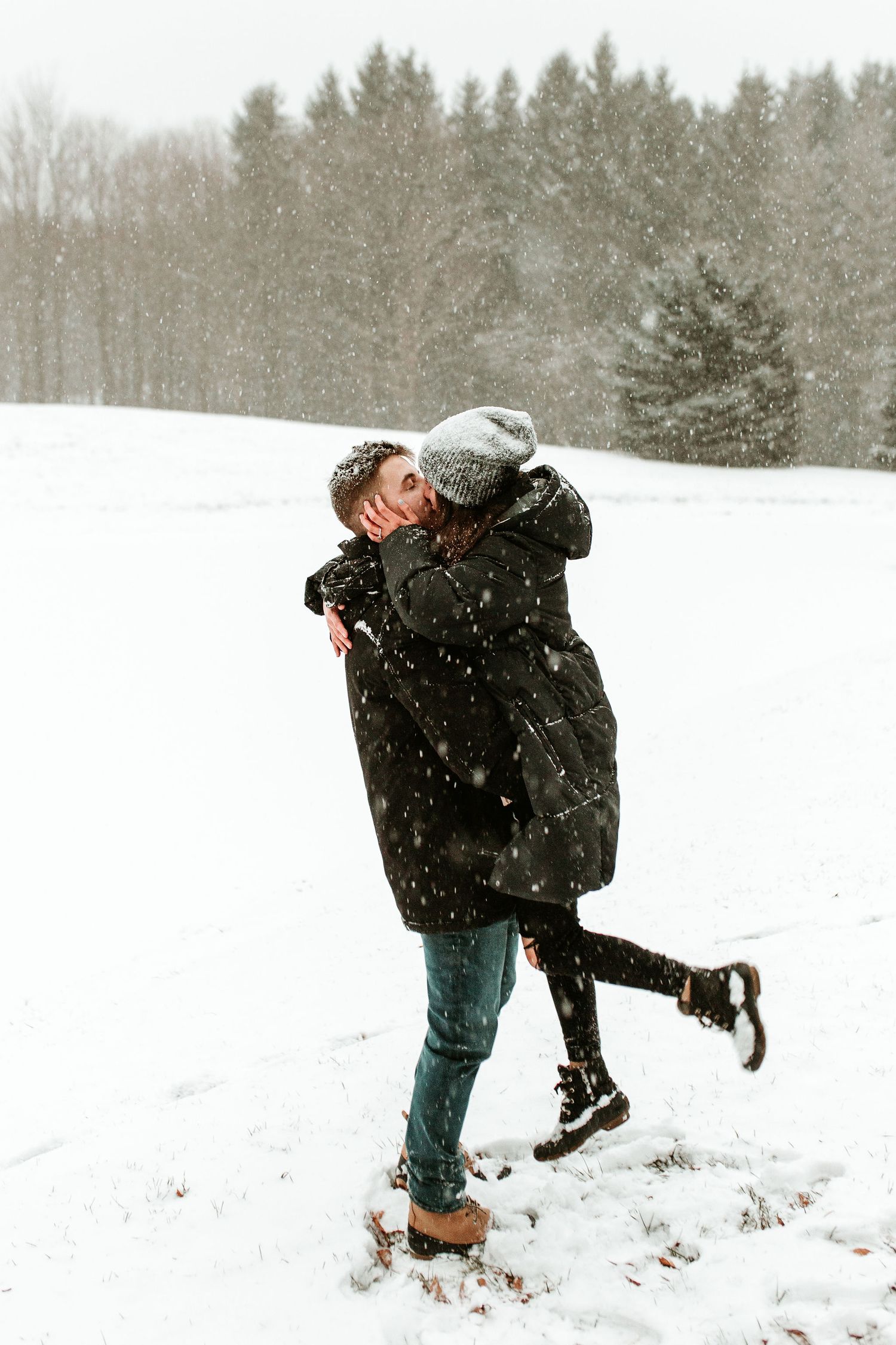 Couple sharing a romantic kiss in a snowy winter landscape, wearing dark coats and a gray beanie hat.