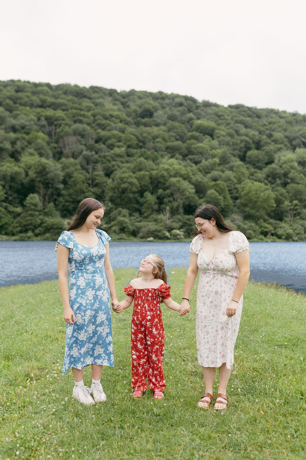 Three people in summer dresses walk together through a green meadow with a lake in the background.