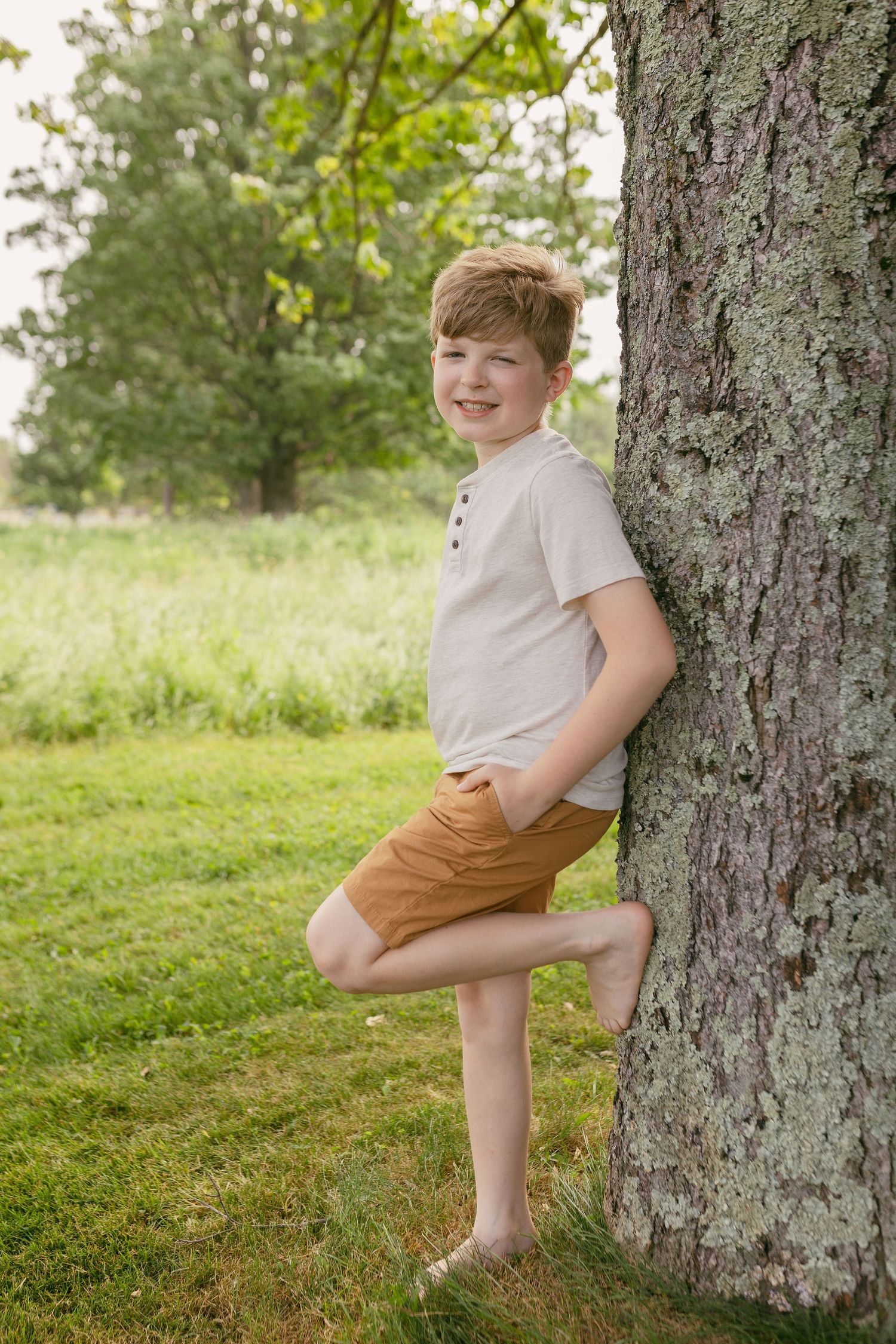 A young child in a white shirt and brown shorts leans against a large tree trunk in a sunny meadow.