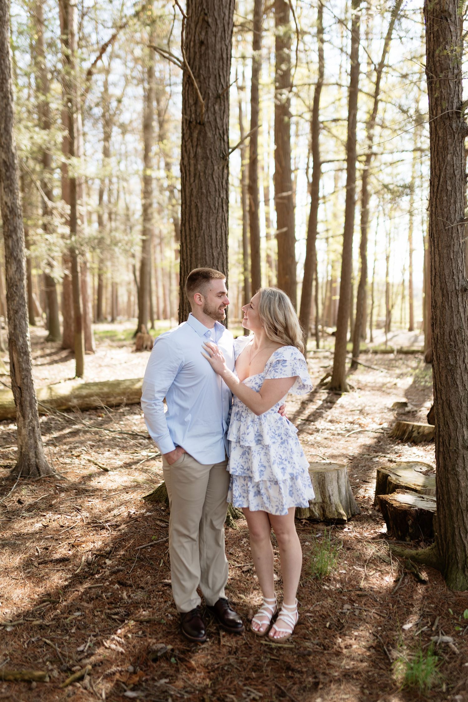 Couple smiling at each other in a sunlit pine forest, woman in floral dress, man in light blue shirt.