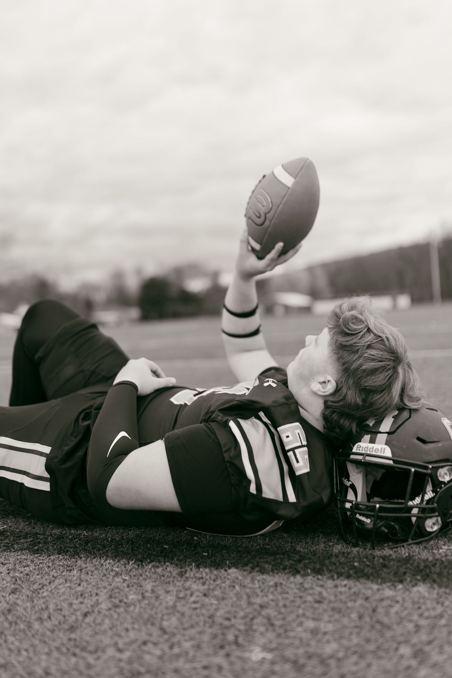 Black and white photo of a football player lying on field, holding ball up while resting head on helmet.