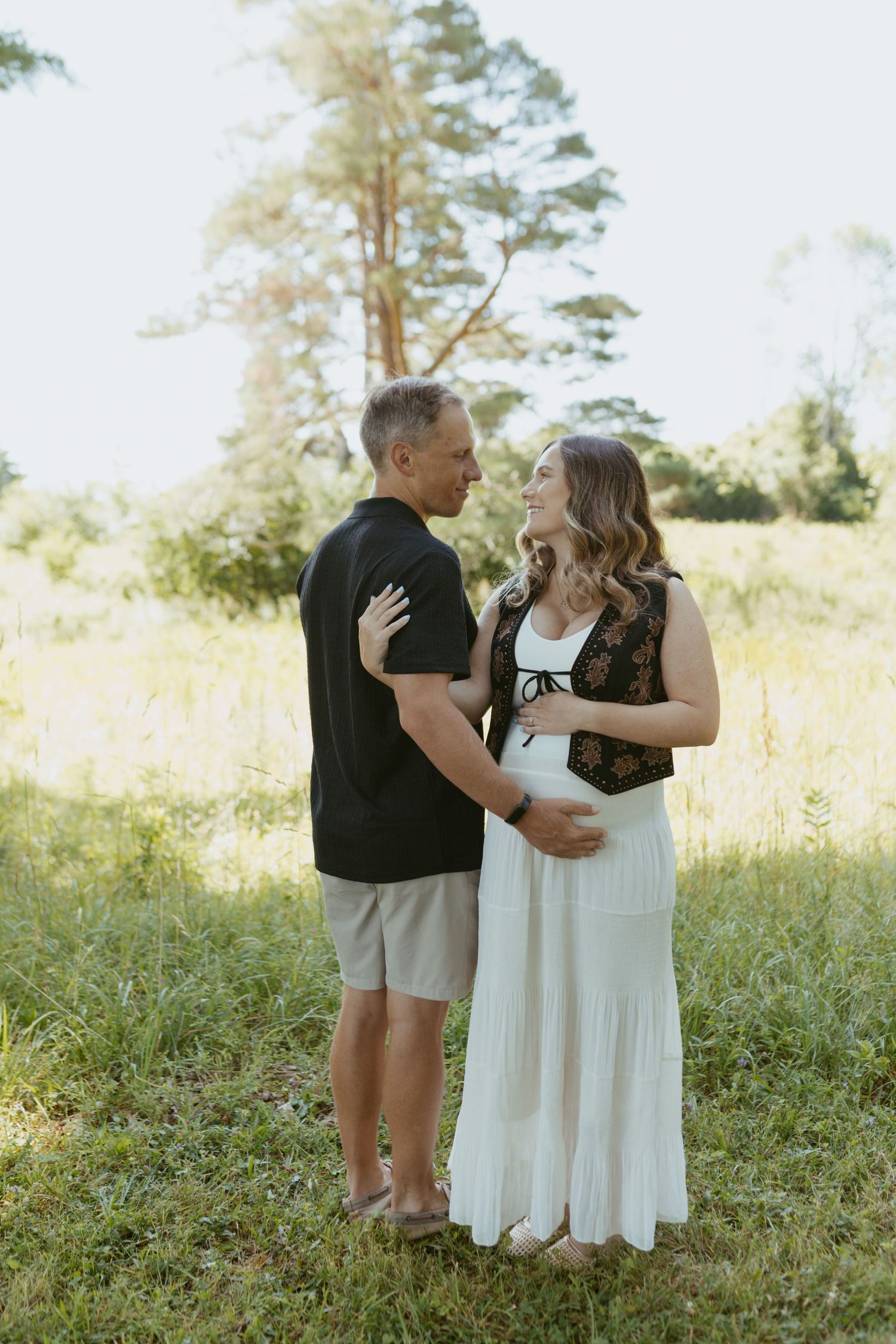 Couple sharing a tender moment outdoors, pregnant woman in white dress embracing man in black shirt in a sunny field.