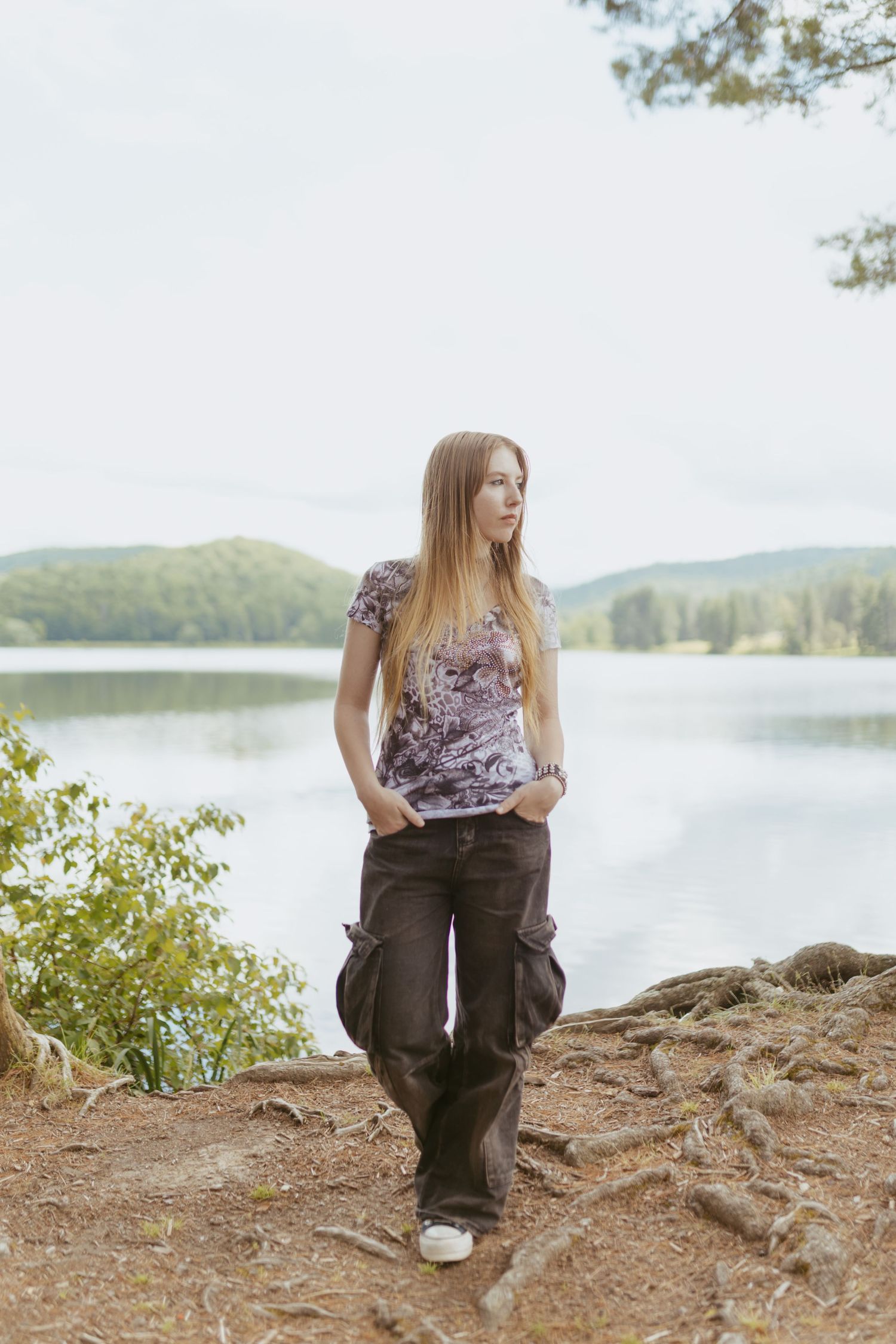 Young woman in cargo pants and floral top standing by a serene lake surrounded by lush green trees.