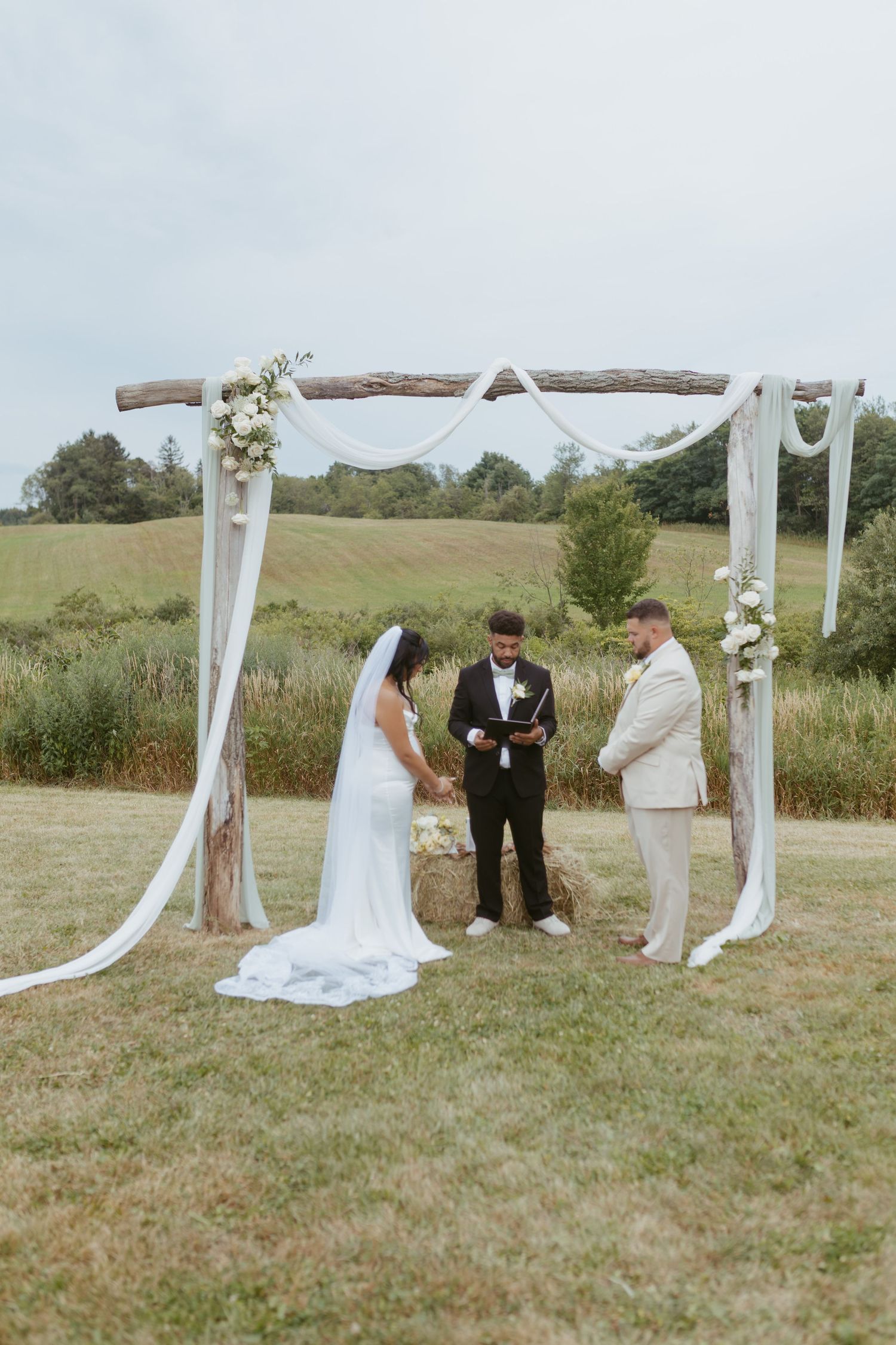 Bride and groom exchange vows under a rustic wooden arch draped with white fabric and flowers in an outdoor field.