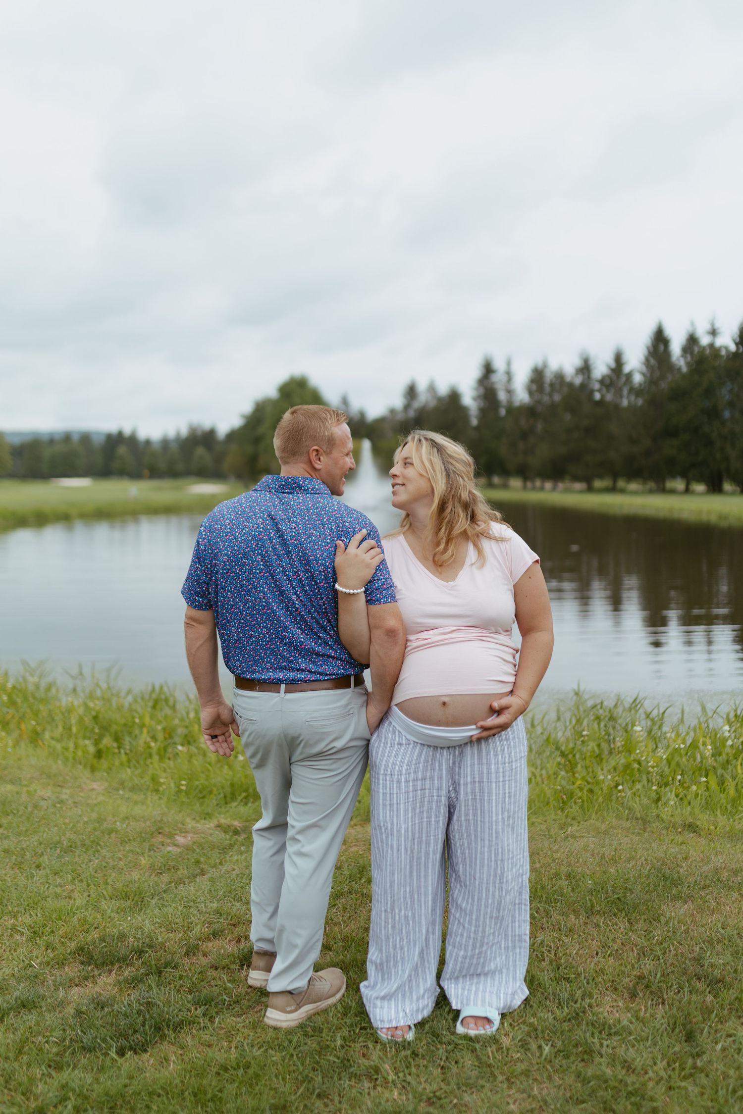 Pregnant couple standing by a serene lake, smiling at each other on a lush green lawn with trees in background.