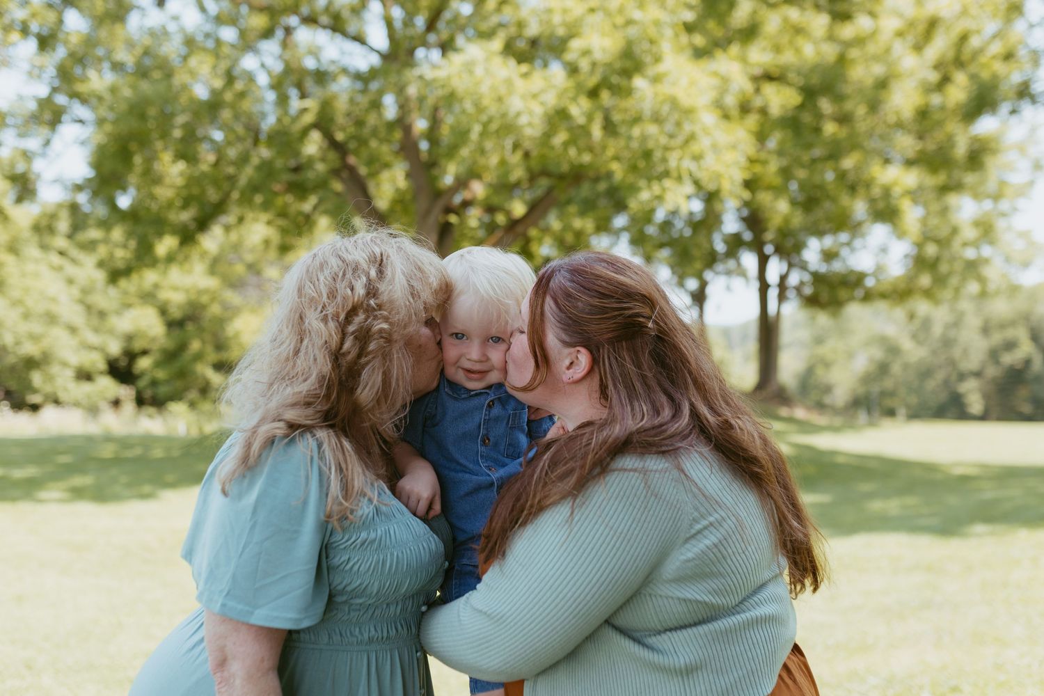 Two women kiss the cheeks of a smiling toddler in a sunny green park, capturing a joyful family moment.