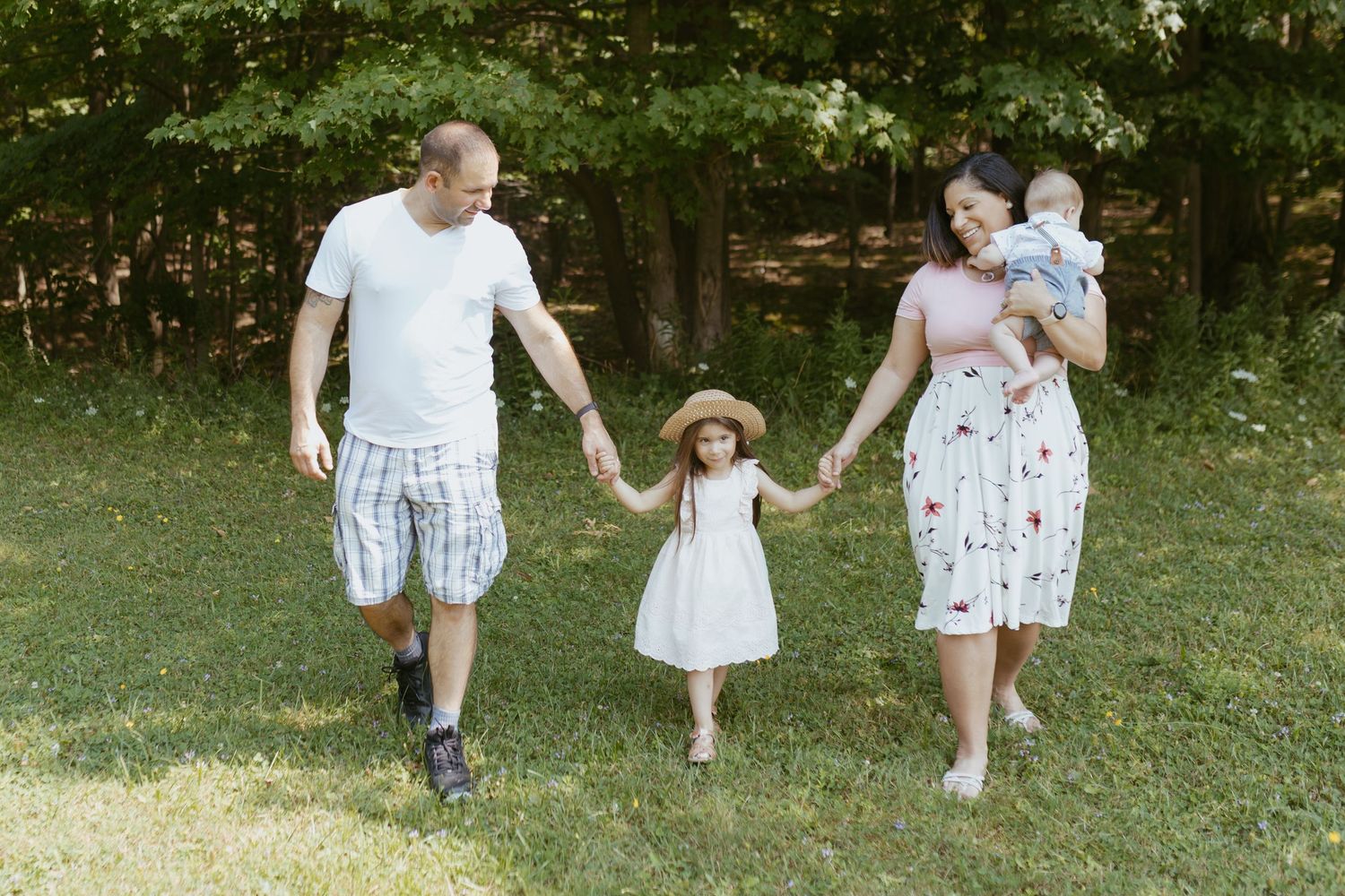 Family of four walking hand-in-hand outdoors, parents with toddler girl in hat and baby, on green grass near trees.
