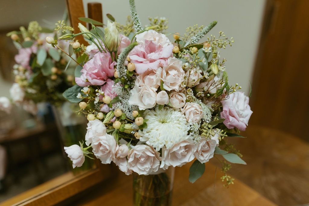 Elegant bridal bouquet featuring soft pink roses, white chrysanthemums, and greenery arranged in a glass vase.