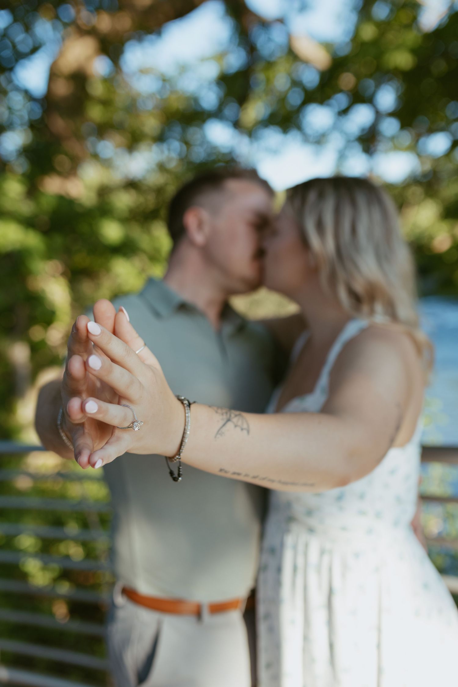 Couple kissing outdoors while showing off engagement ring, woman wearing floral dress with bracelet and tattoo.