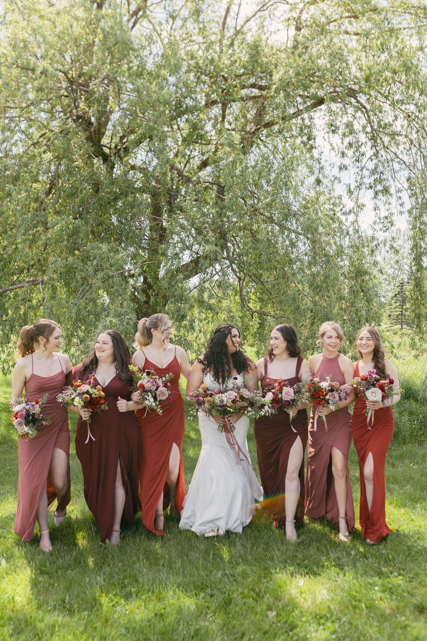 Wedding party in burgundy dresses posed under willow trees in natural outdoor setting.