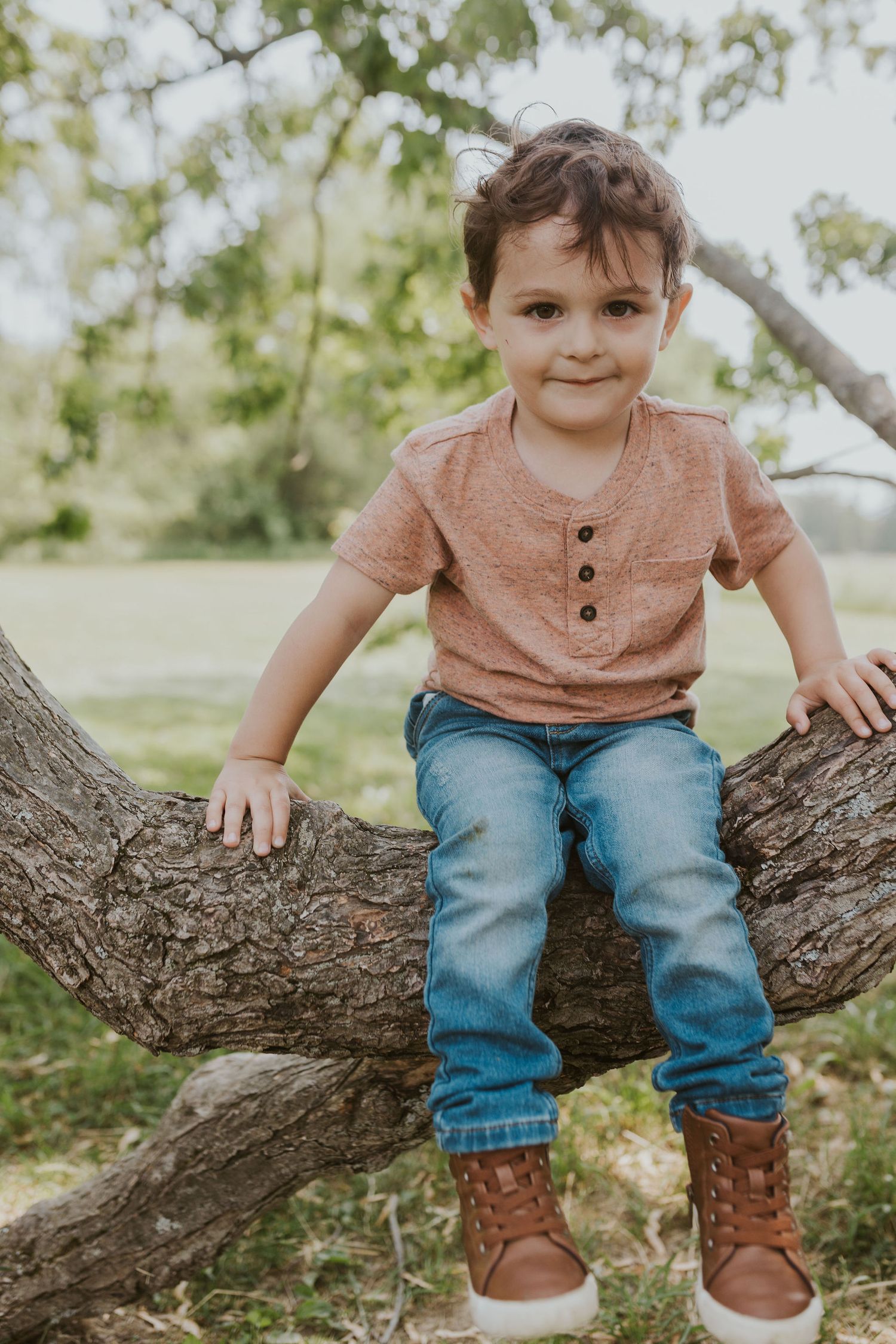 Small child sitting on a tree branch wearing a coral shirt and jeans with brown boots.