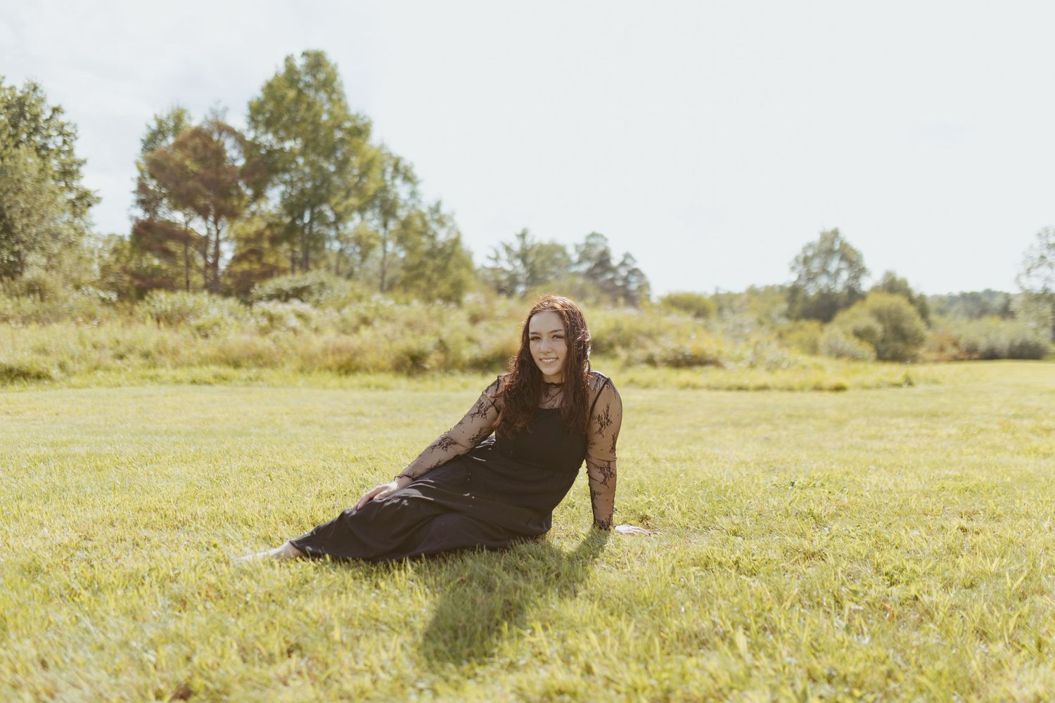 Woman in black dress sitting on green grass field with trees in background on a sunny day.