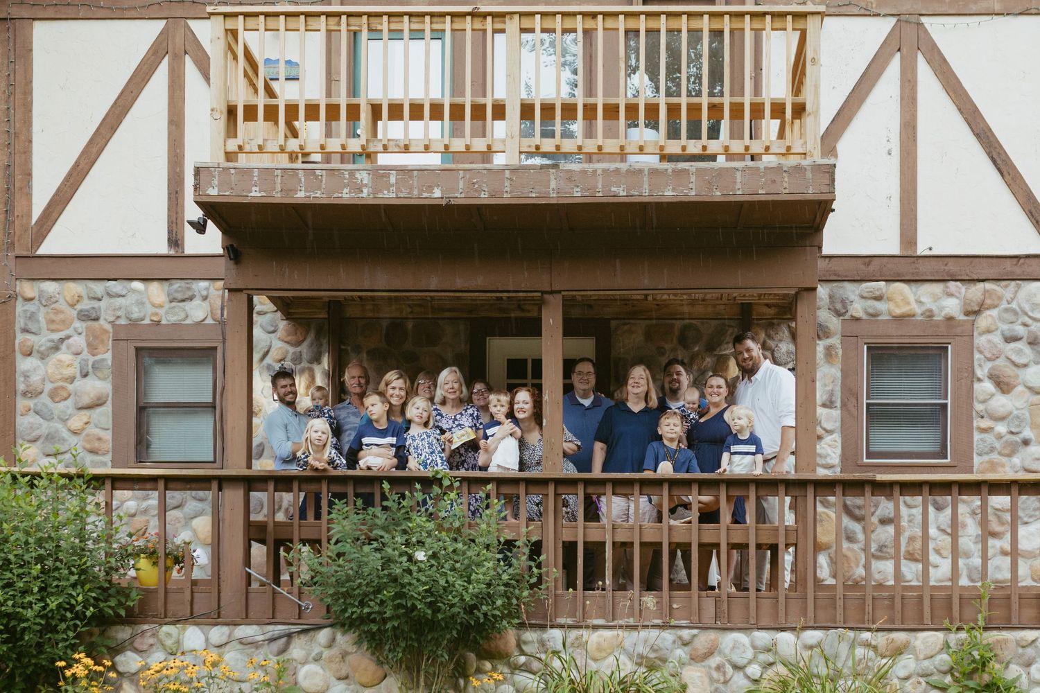 Large family group posing on a stone building porch with upper balcony, smiling together outdoors.