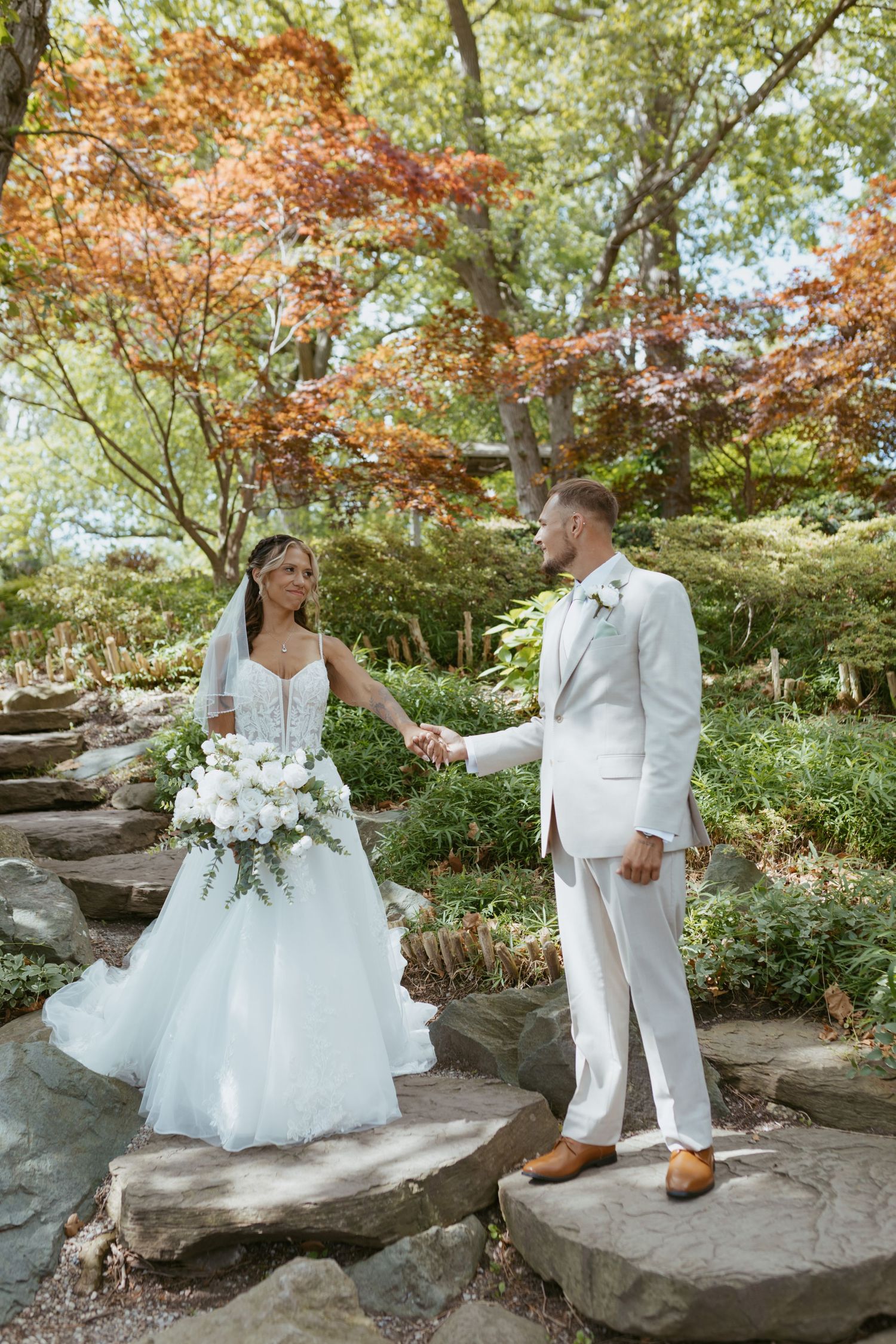 A bride and groom hold hands on stone steps in a lush garden, she in a white gown with bouquet, he in a white suit.