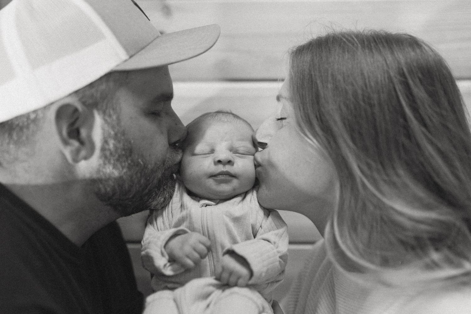 Black and white photo of two parents kissing their newborn baby on the cheeks in a tender family moment.