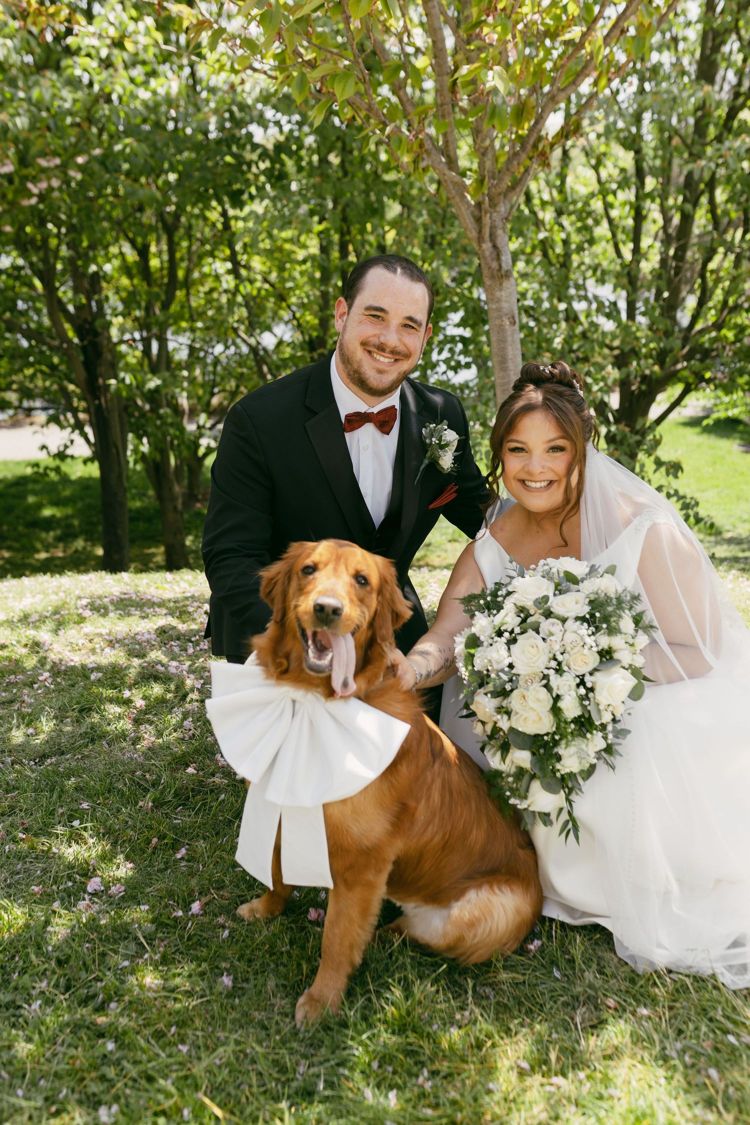 Bride and groom pose with their golden retriever wearing a white bow collar on their wedding day outdoors.
