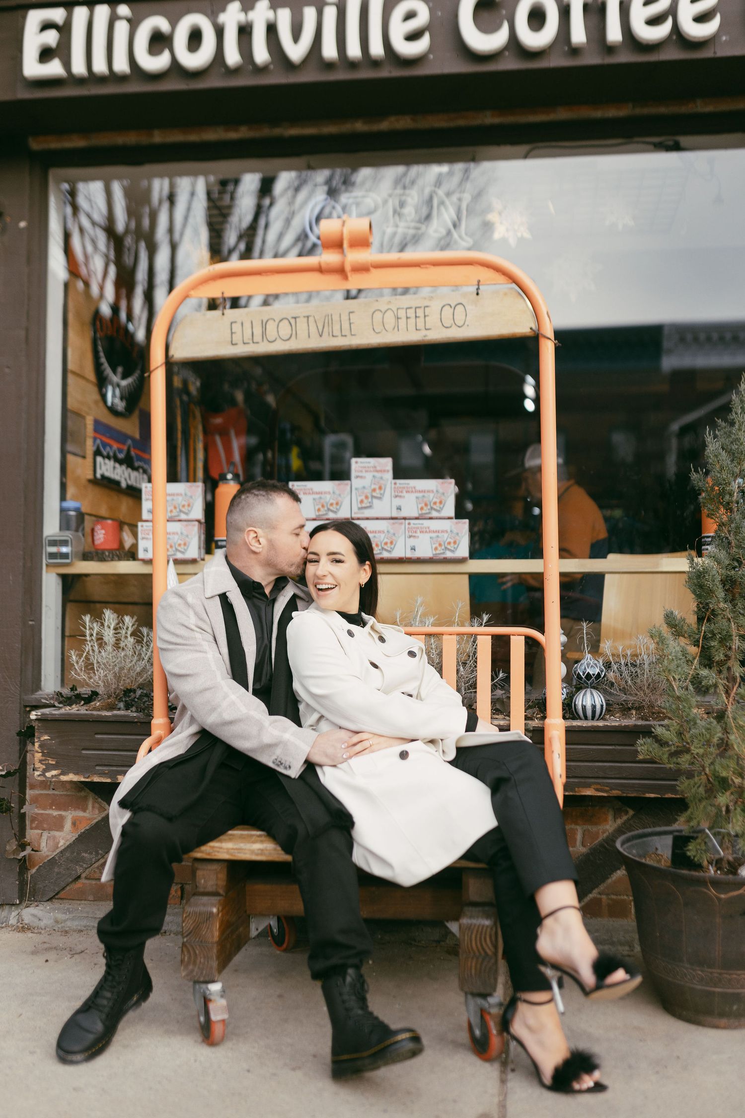 Couple sitting on bench outside Ellicottville Coffee Co, sharing a romantic kiss on a cool day.