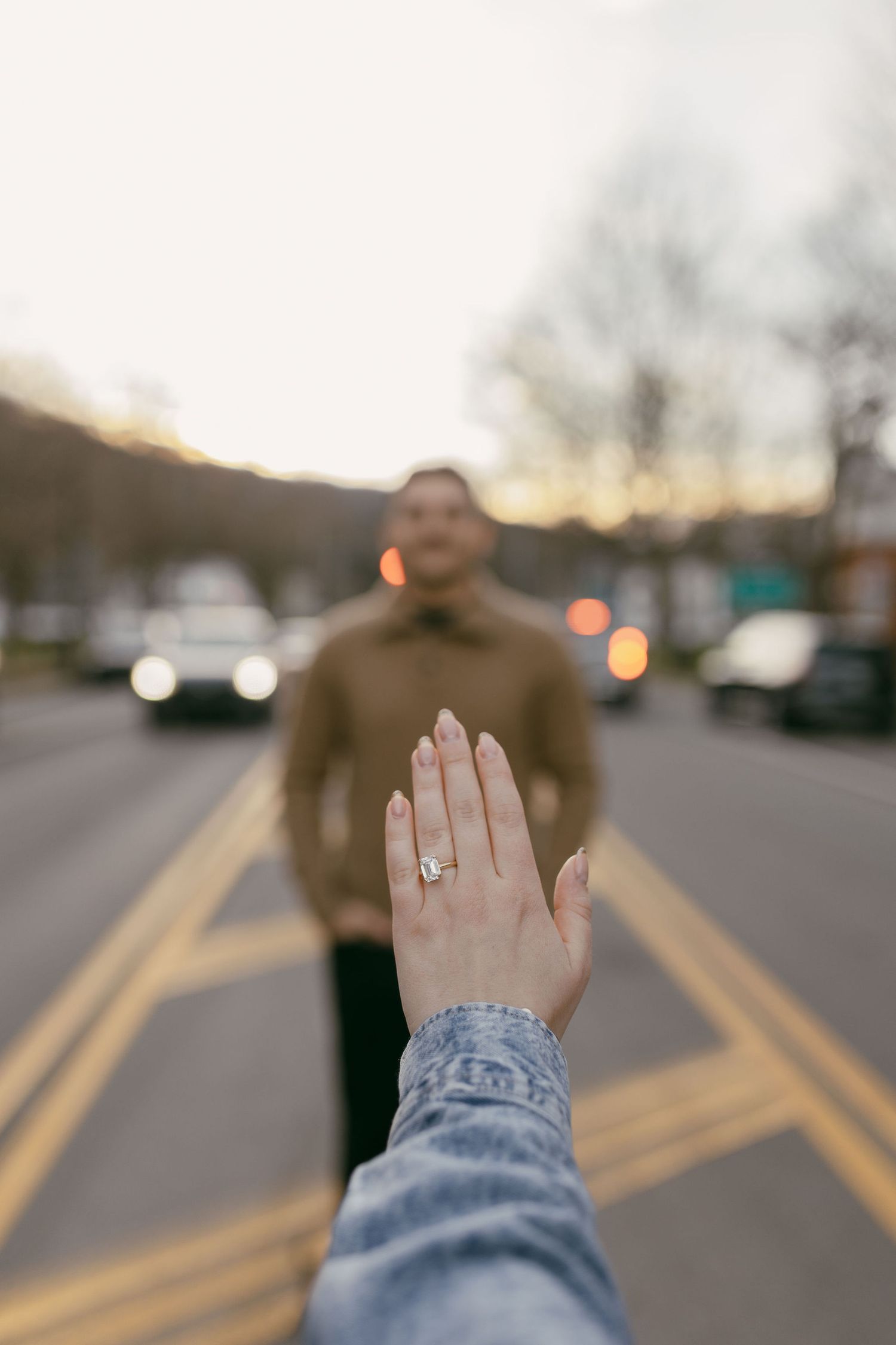 Close-up of an engagement ring on a woman's hand reaching toward a smiling man standing on a street at dusk.