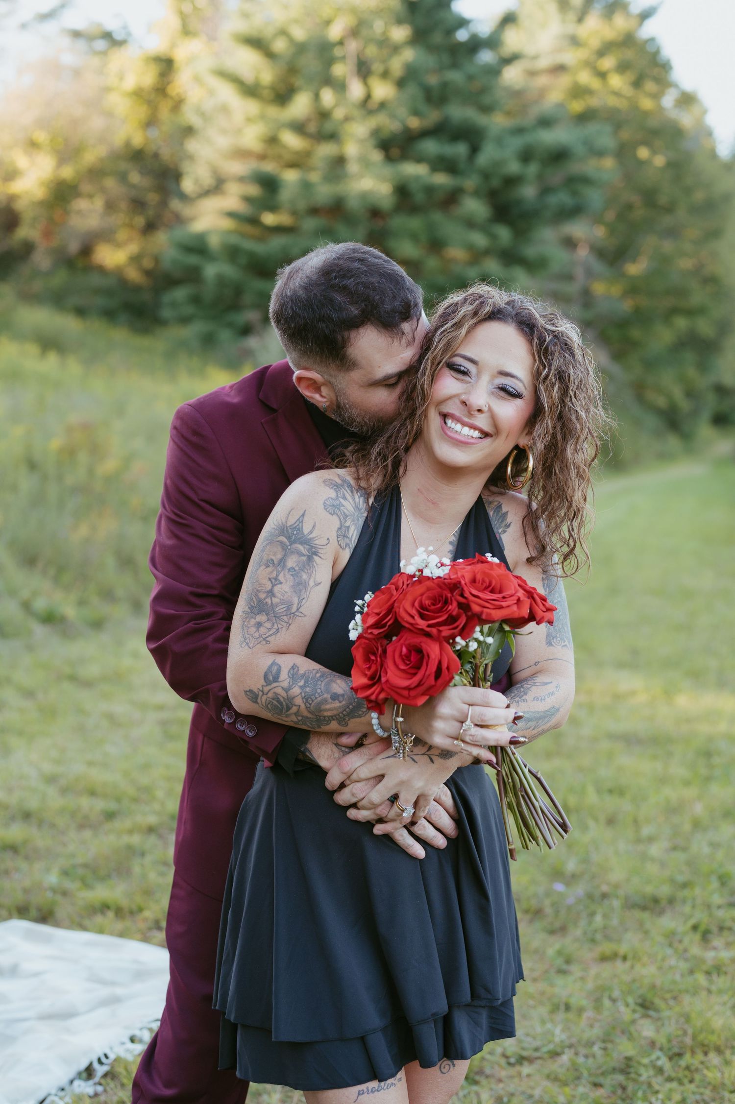 A smiling woman in a black dress holds red roses while a man in a burgundy suit kisses her cheek outdoors.
