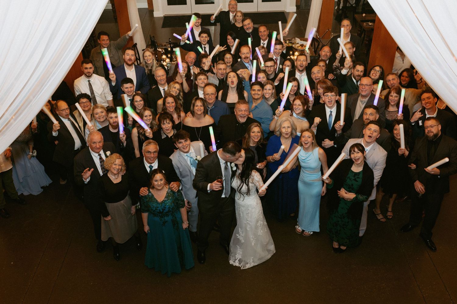 Wedding guests dance and celebrate on a lively dance floor with colorful lighting and white drapes in the background.