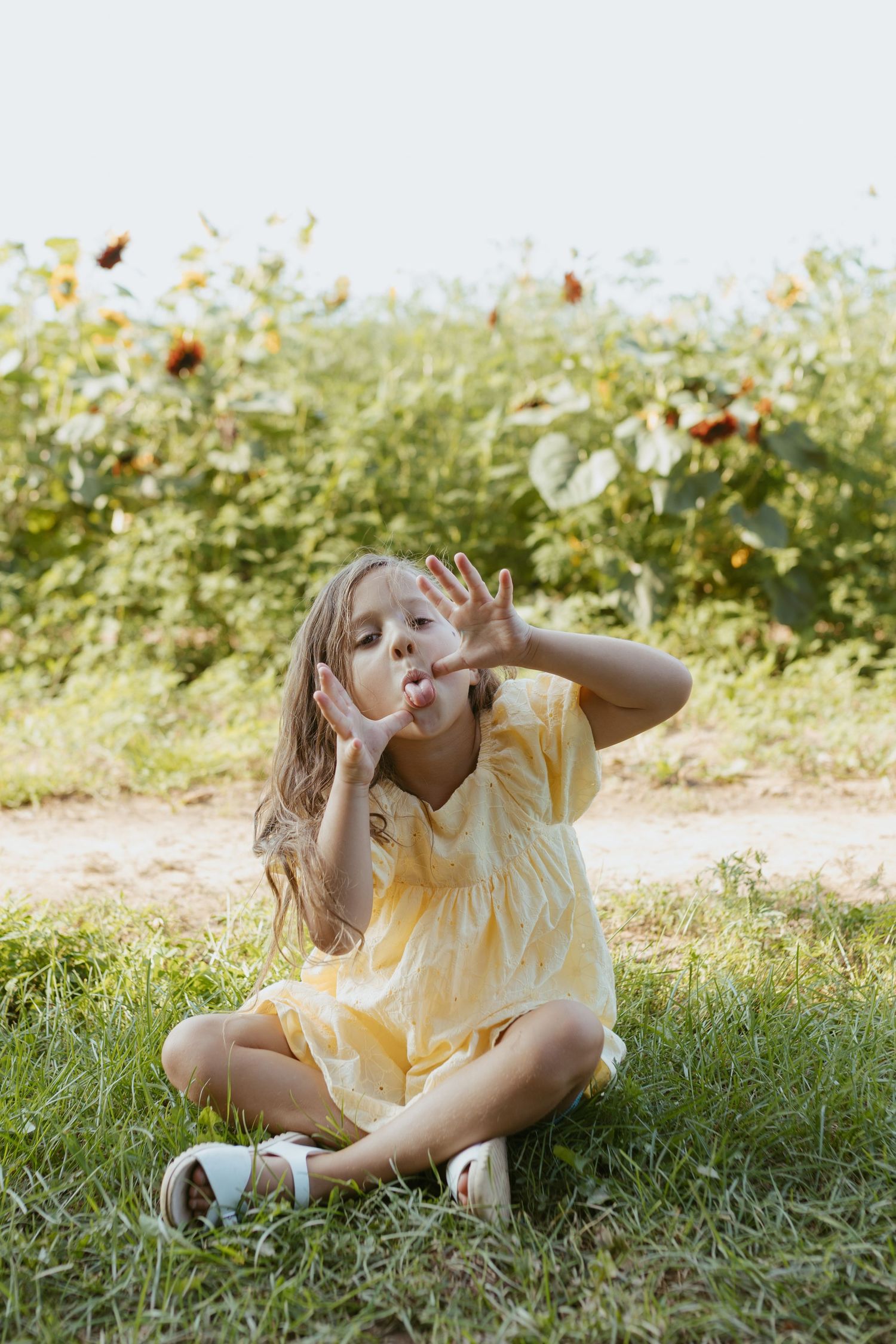 Young girl in yellow dress sitting cross-legged on grass, blowing a kiss with hands raised, sunflowers in background.