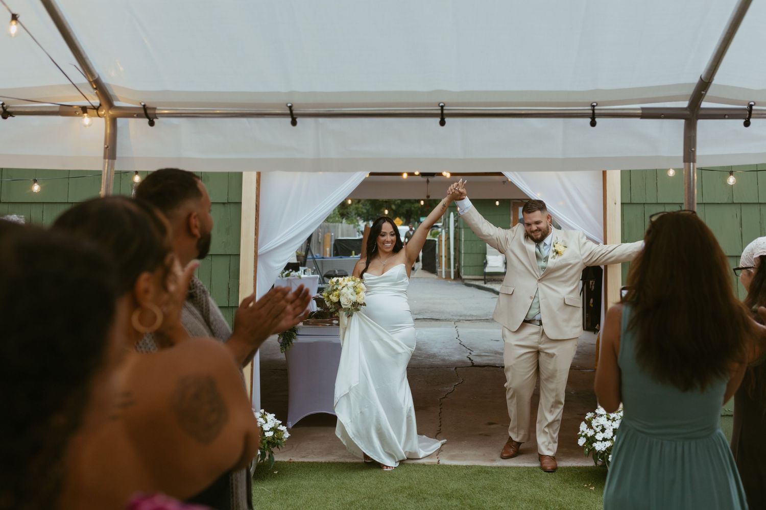 Bride and groom raise hands joyfully under a white tent as guests celebrate their wedding reception entrance.