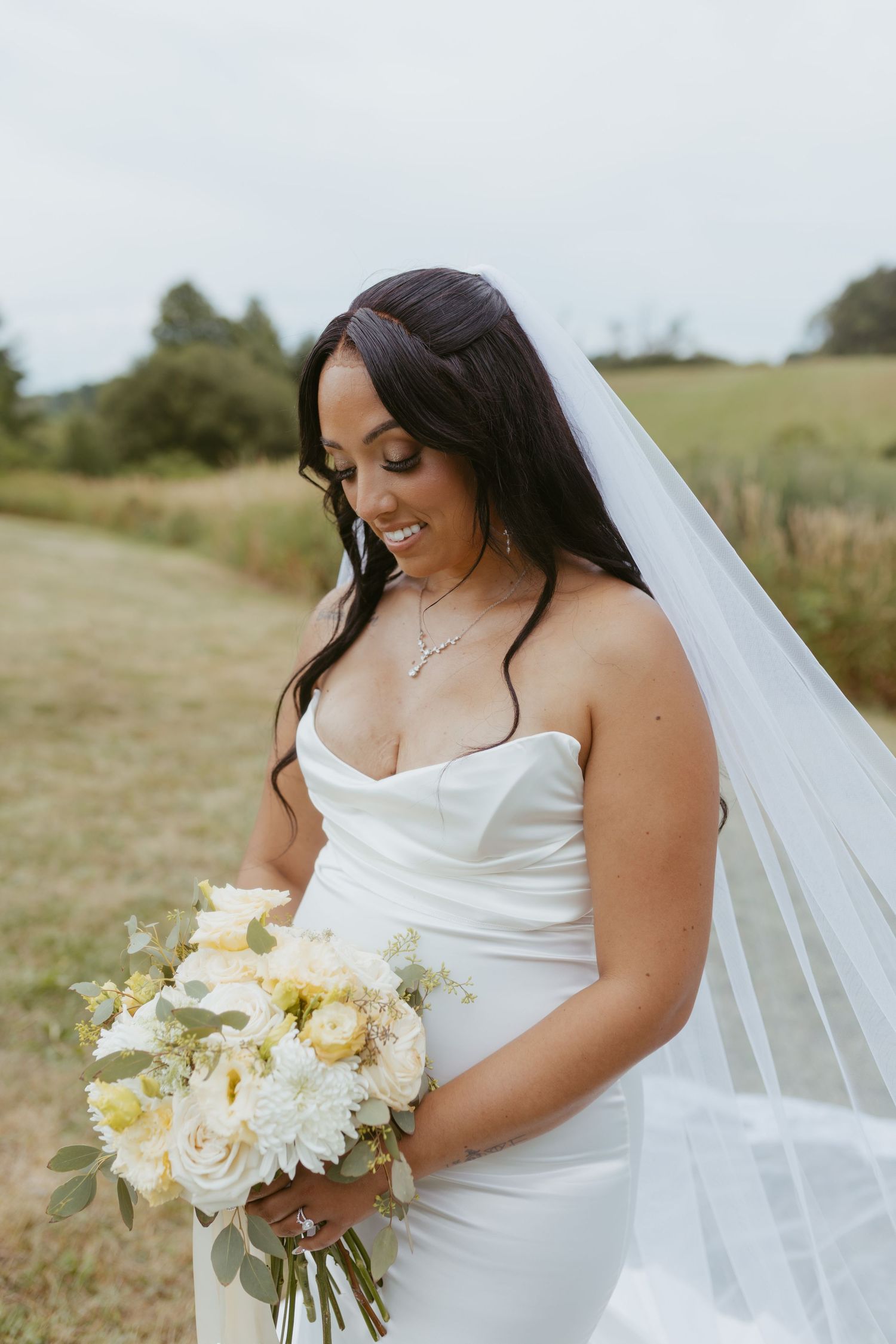Smiling bride in strapless white gown and veil holds white bouquet outdoors in green field on wedding day.