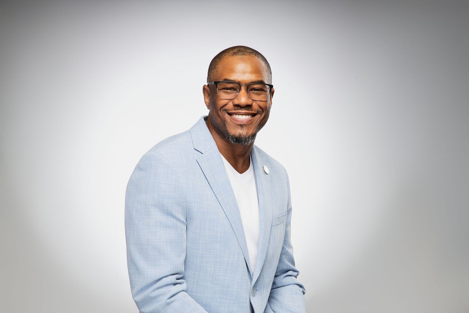 Smiling Black man with glasses and goatee wearing a light blue blazer and white shirt against a gray background.