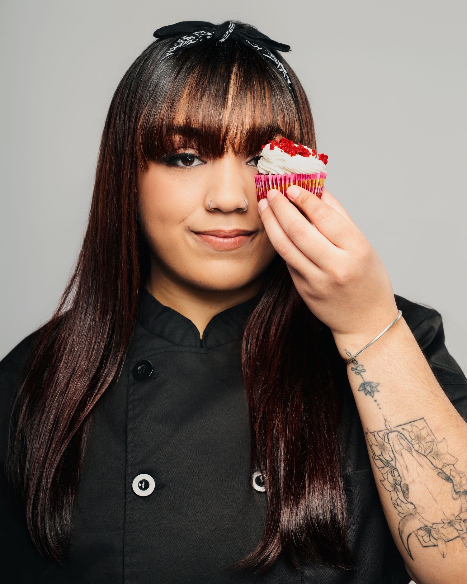 Chef in black uniform holds a pink frosted cupcake while posing against a gray background.