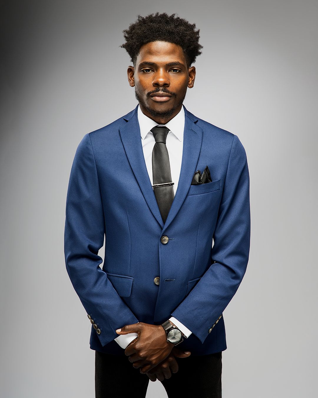 Professional headshot of man in navy blue suit and black tie against grey background.