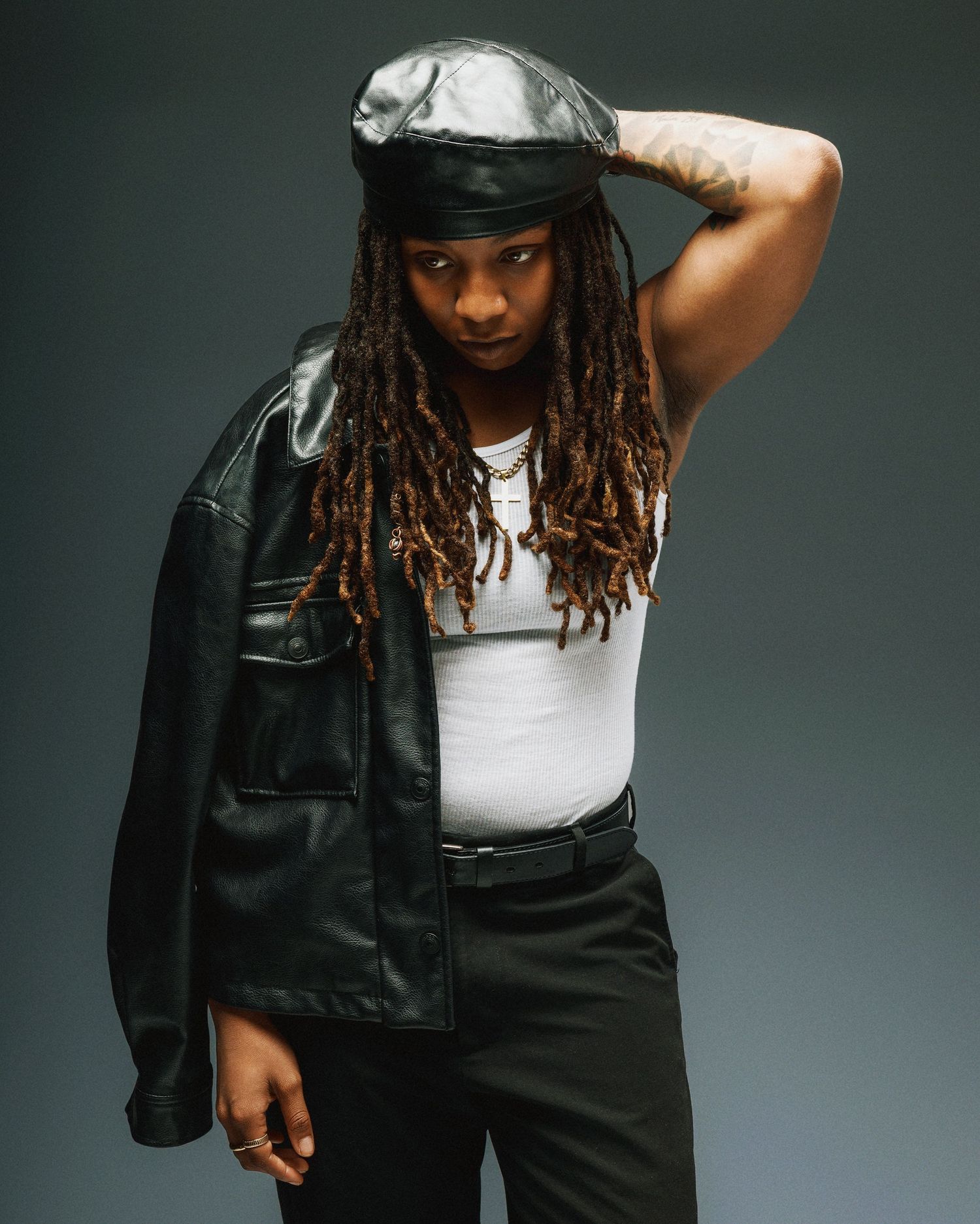 A fashion model poses in a white tank top and black jacket against a dark studio backdrop.