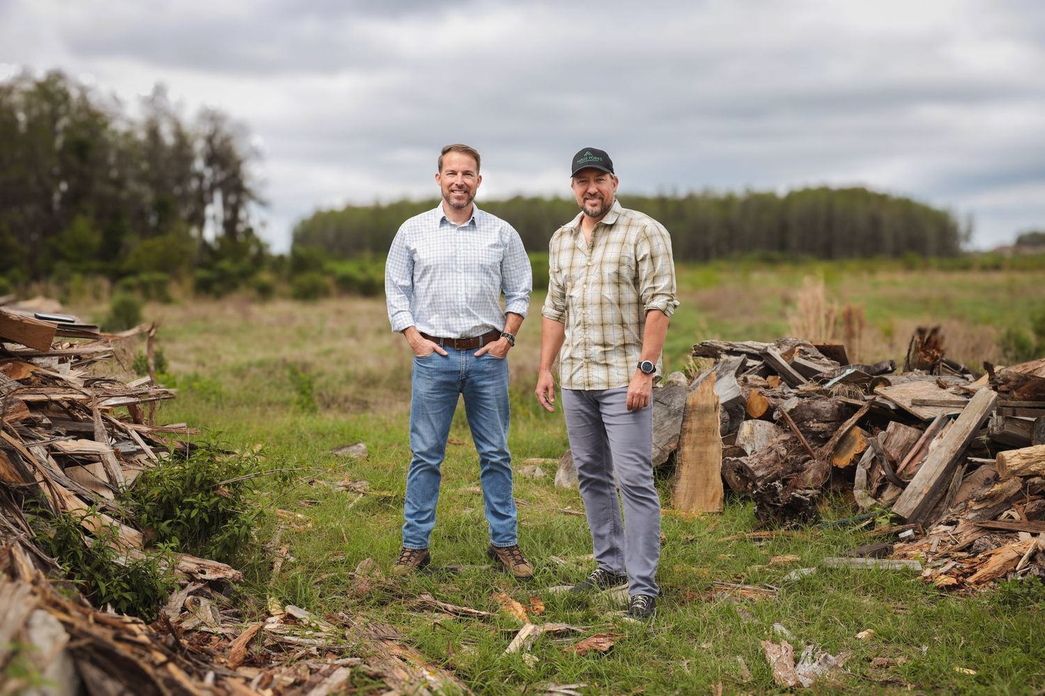 Two people stand in a grassy field next to piles of firewood on a cloudy day.