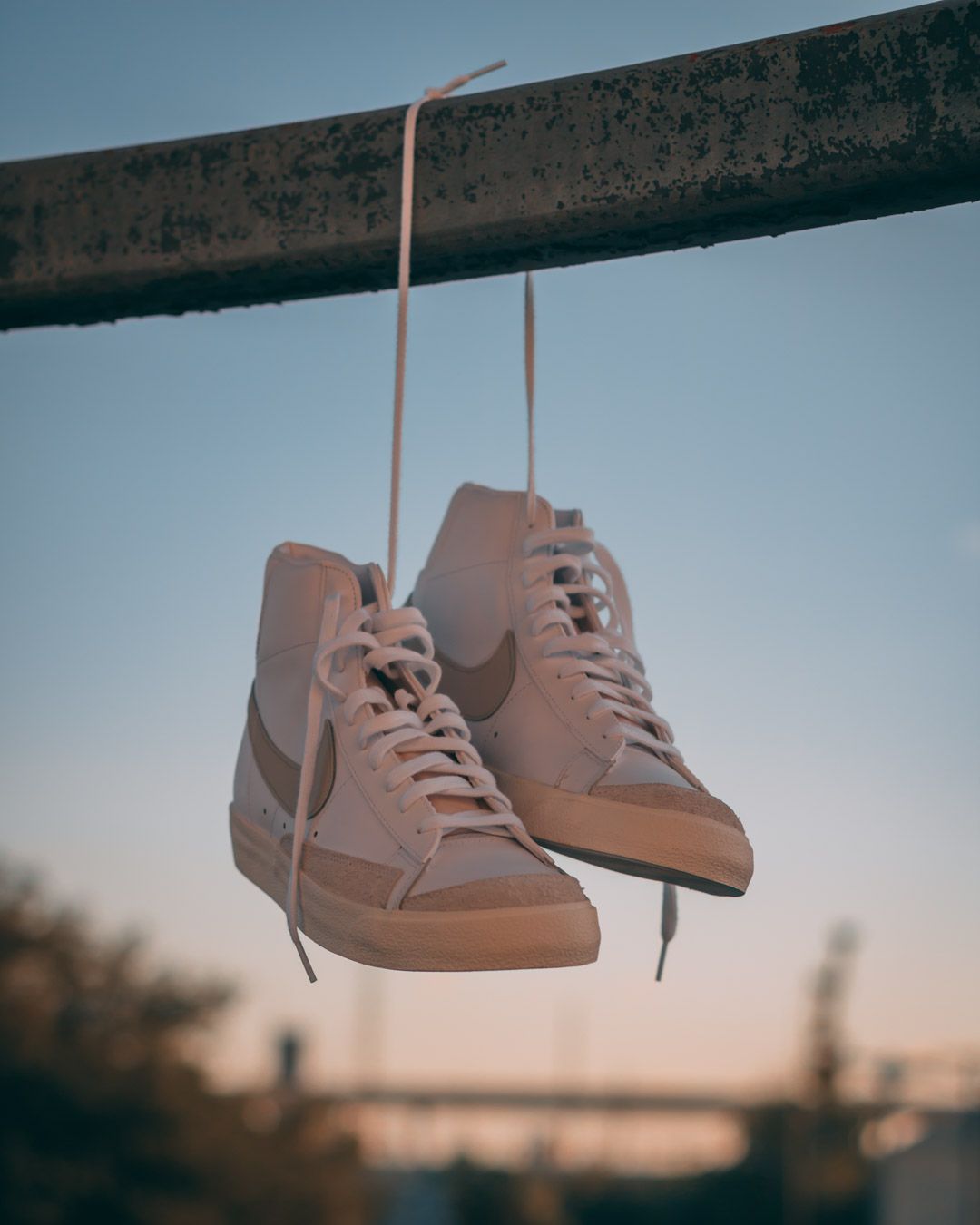 A pair of beige high-top sneakers suspended by laces against a blurred sunset sky background.