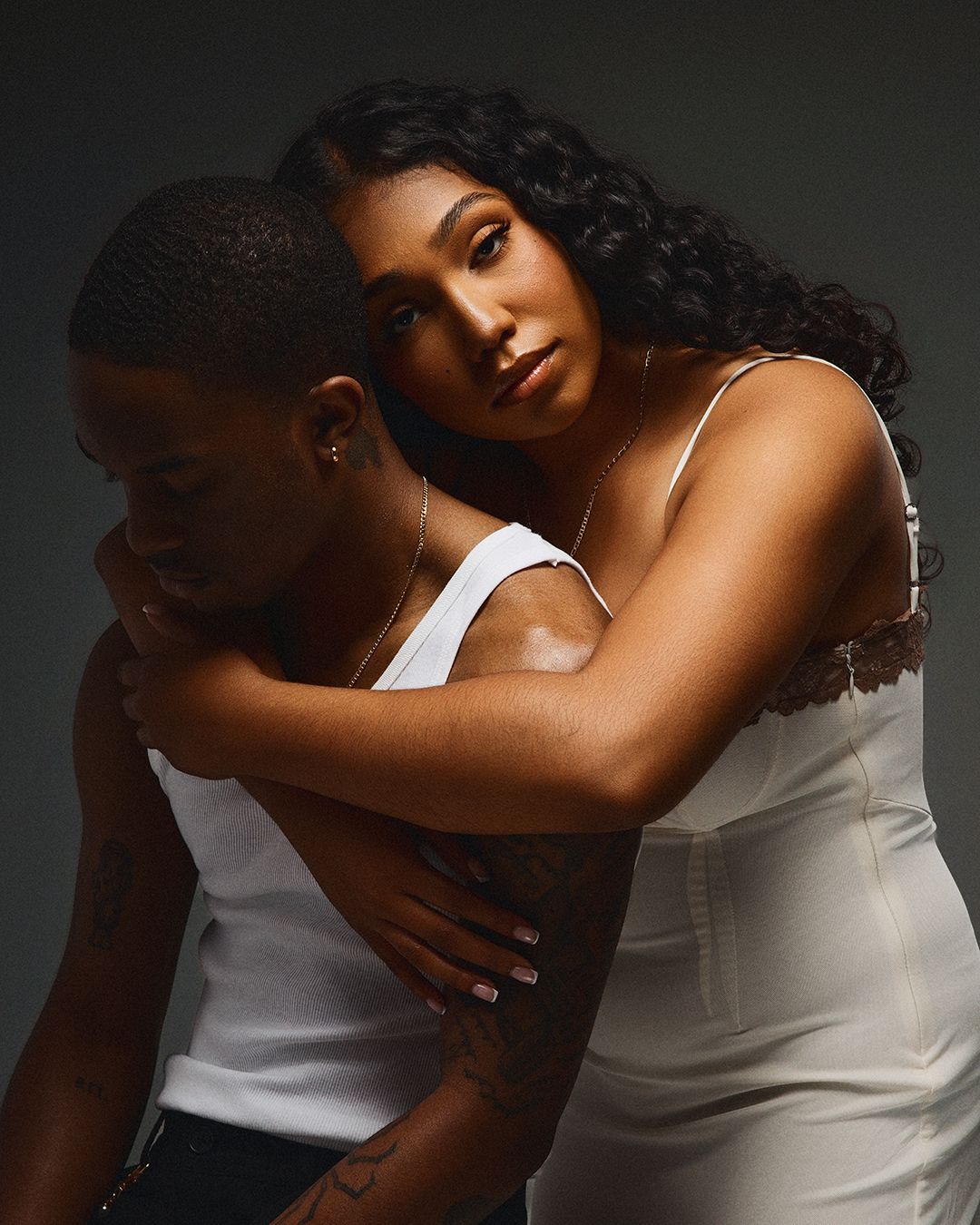 A couple in white outfits share an intimate embrace against a dark studio background.