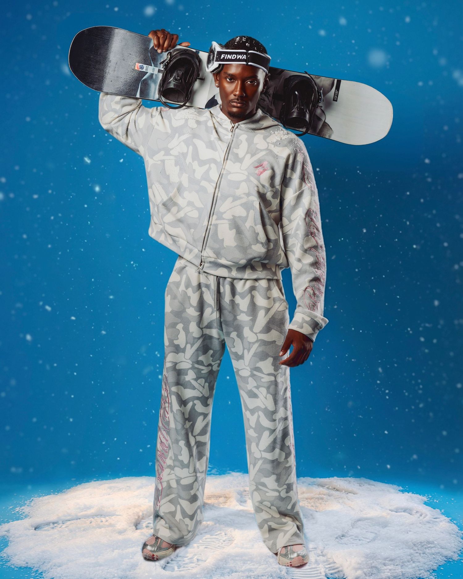 A person in a camouflage snowsuit holds a snowboard against a snowy blue background with falling snowflakes.